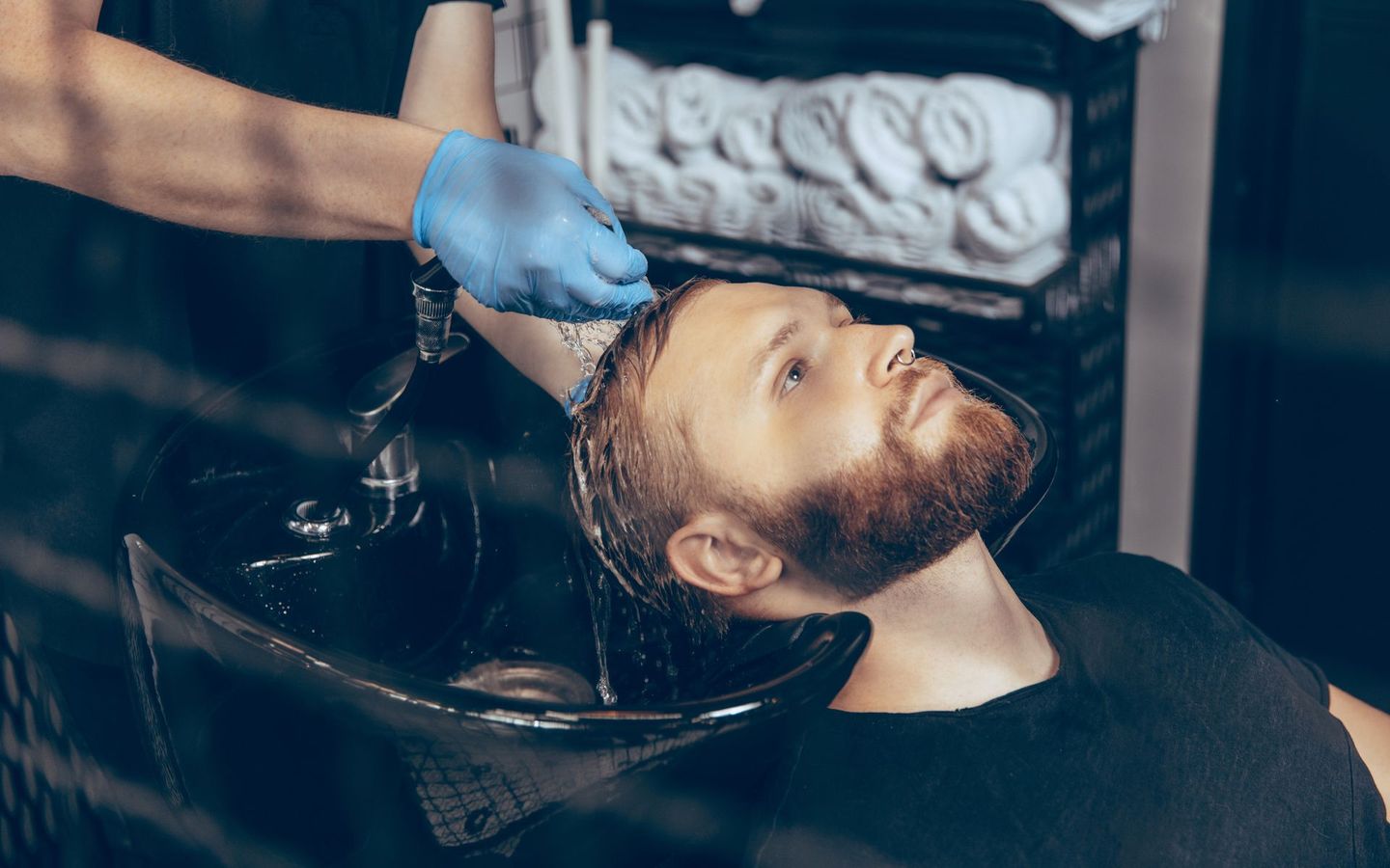Un uomo si fa lavare i capelli in un salone di bellezza; una persona che indossa un guanto blu gli versa dell'acqua sulla testa.