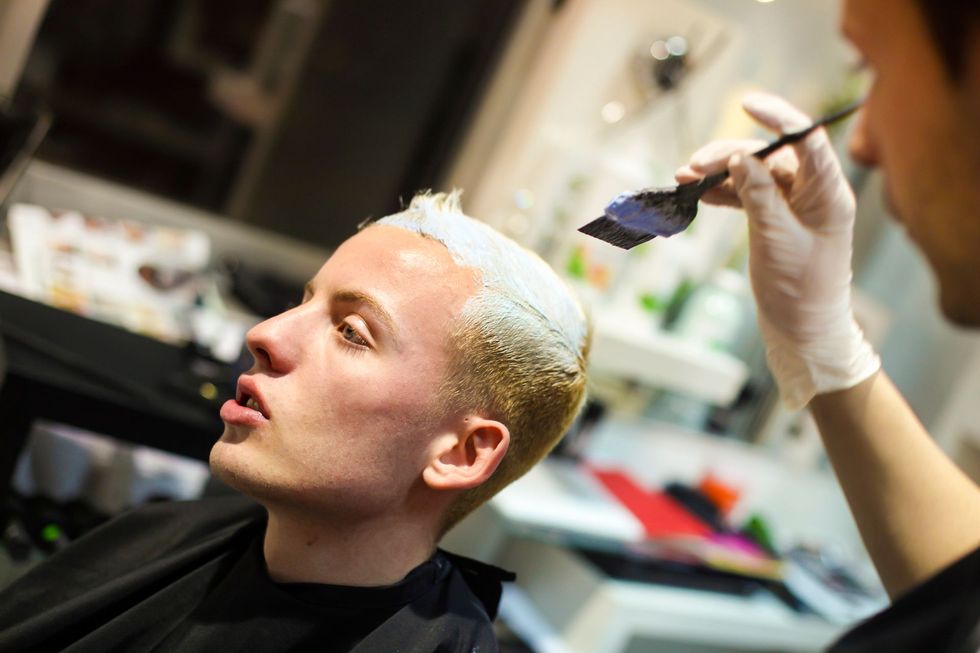 Parrucchiere che tinge i capelli biondi di un uomo in un salone. Lui sembra concentrato; lei indossa i guanti.