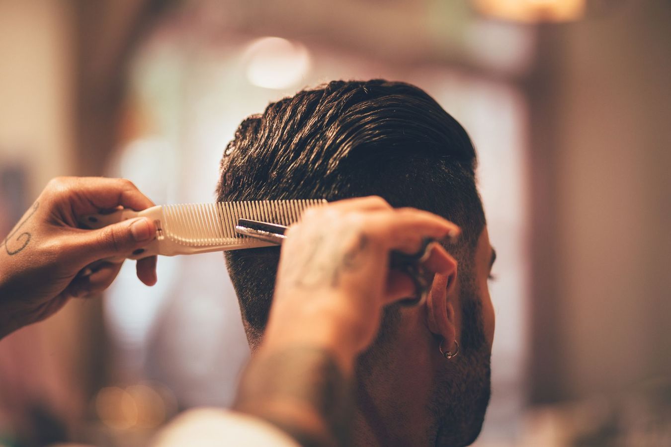 Parrucchiere che pettina i capelli bagnati di un uomo. Mani con tatuaggi, forbici e pettine. Interno, sfondo sfocato.