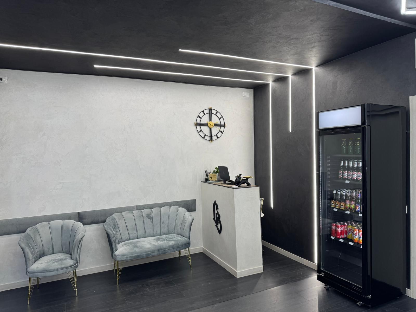 Area reception con divani grigi, bancone bianco, frigorifero per bevande e luci lineari.