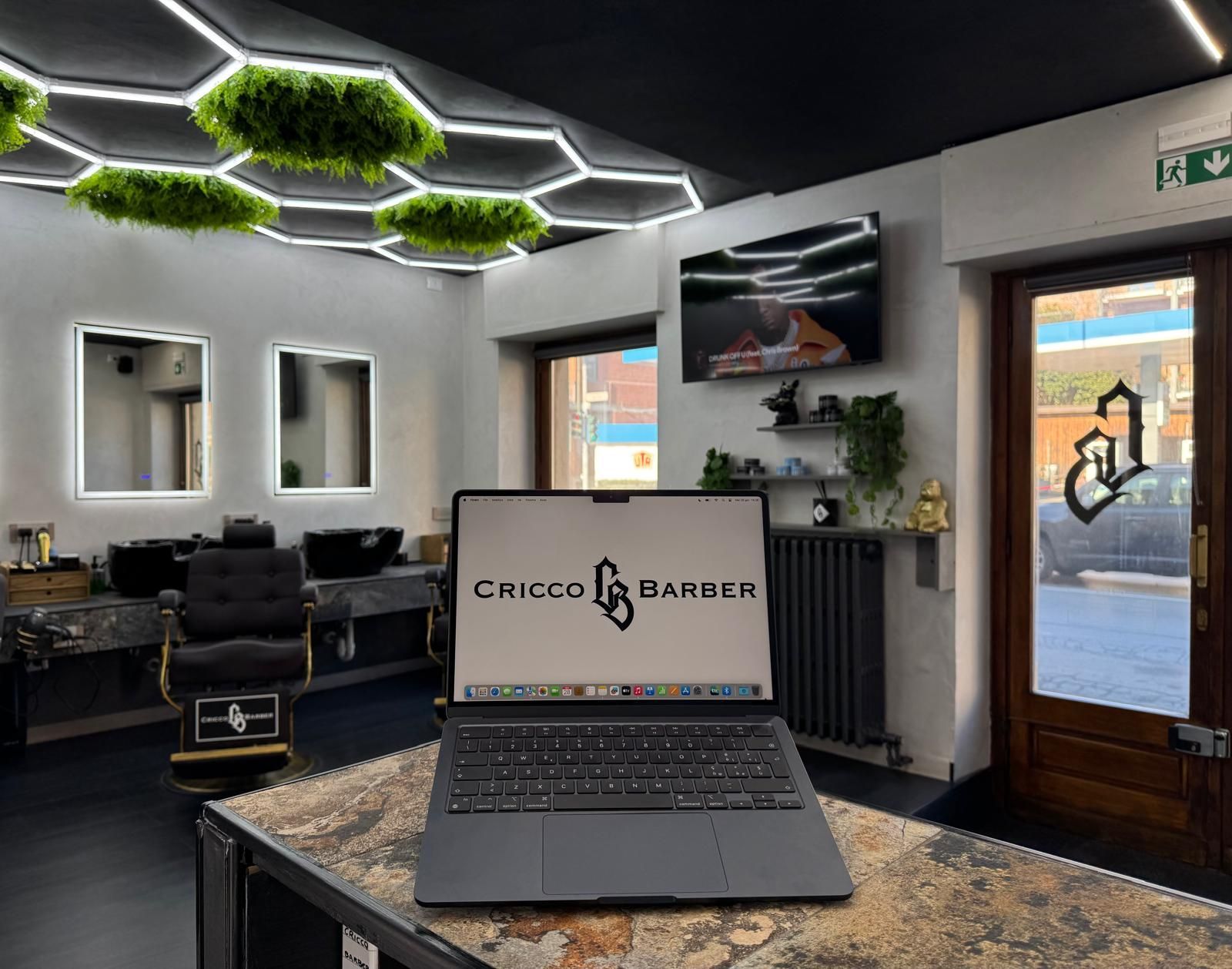 Computer portatile sul bancone di un barbiere con illuminazione decorativa esagonale. Sullo schermo è visibile il logo
