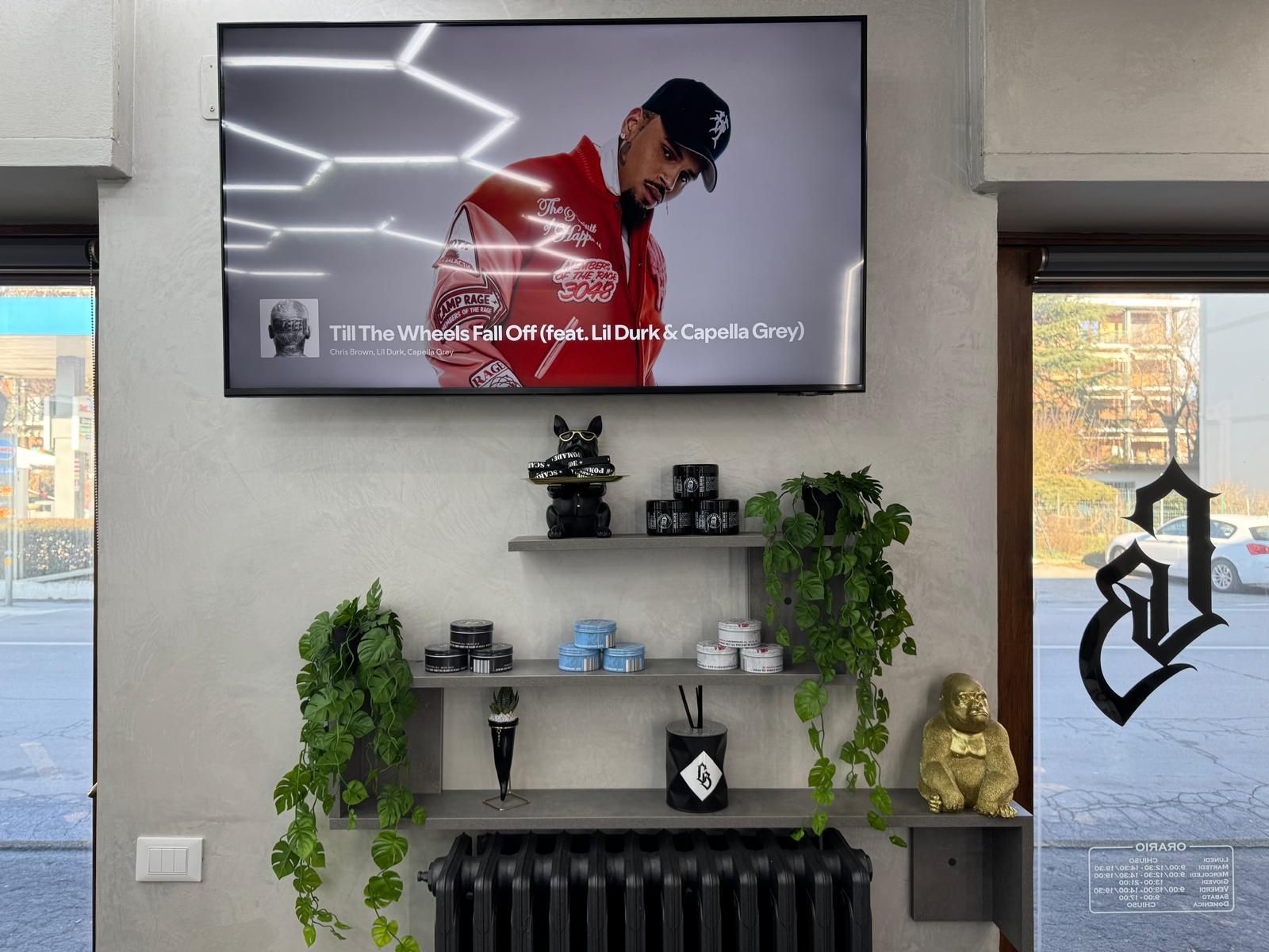 Interno di un barbiere con TV che riproduce video musicali, scaffali con decorazioni e una finestra.