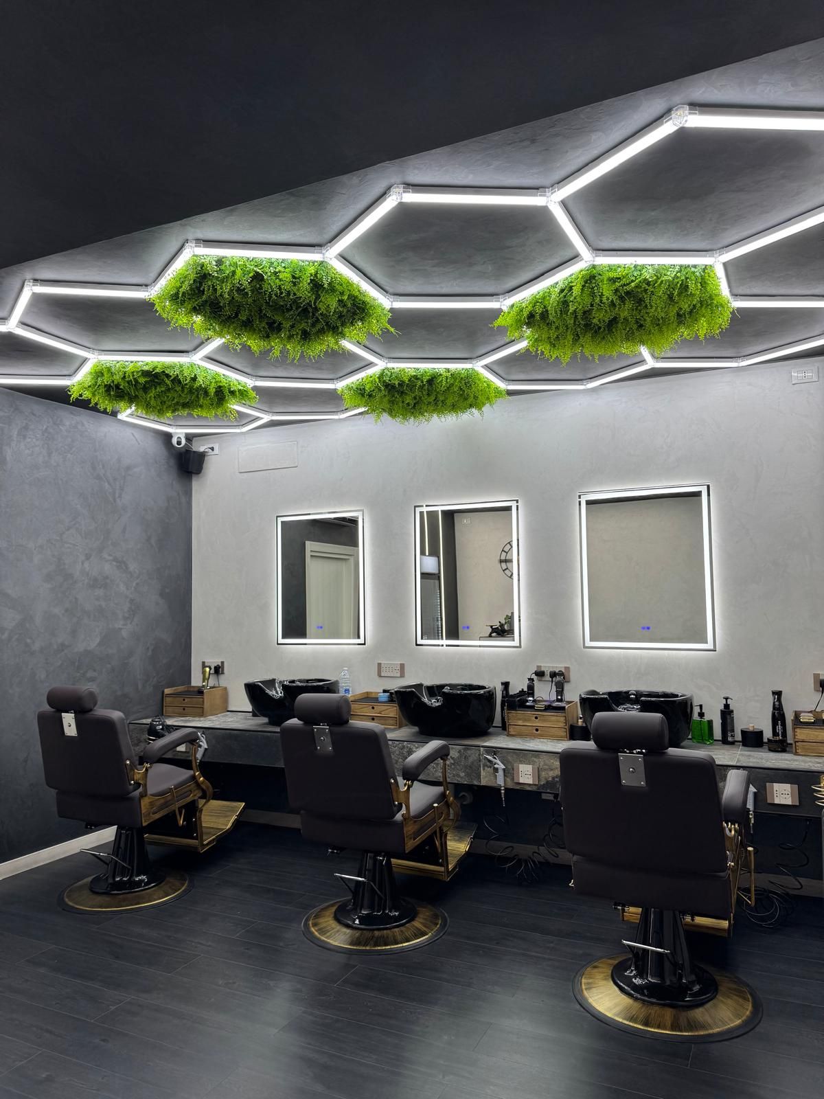 Interno di un salone da parrucchiere con tre sedie, specchi e lampade da soffitto esagonali con muschio.