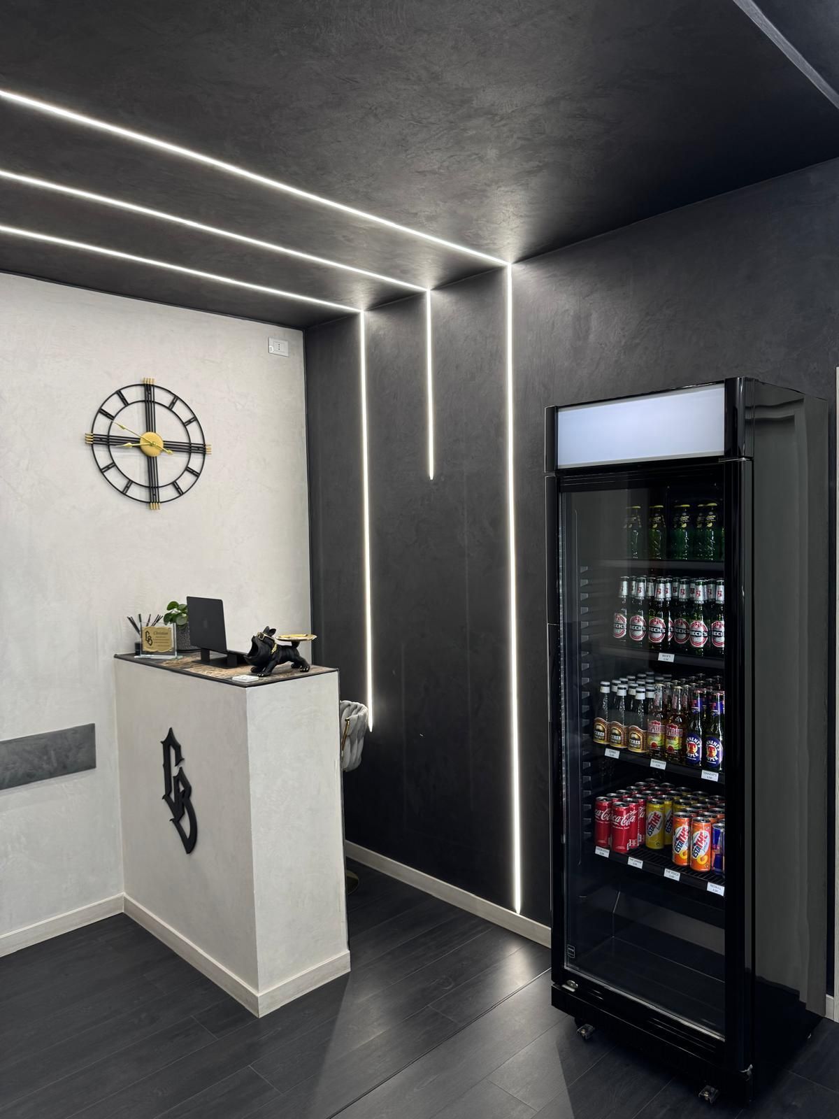 Uno spazio commerciale moderno con bancone, orologio, frigorifero e illuminazione lineare su una parete nera.