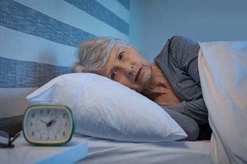 An elderly woman is laying in bed with an alarm clock next to her.