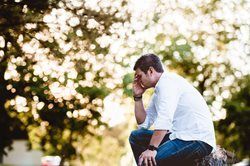 A man is sitting on a rock in a park with his head in his hands.