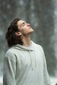 A man in a hoodie is standing in front of a waterfall with his eyes closed.