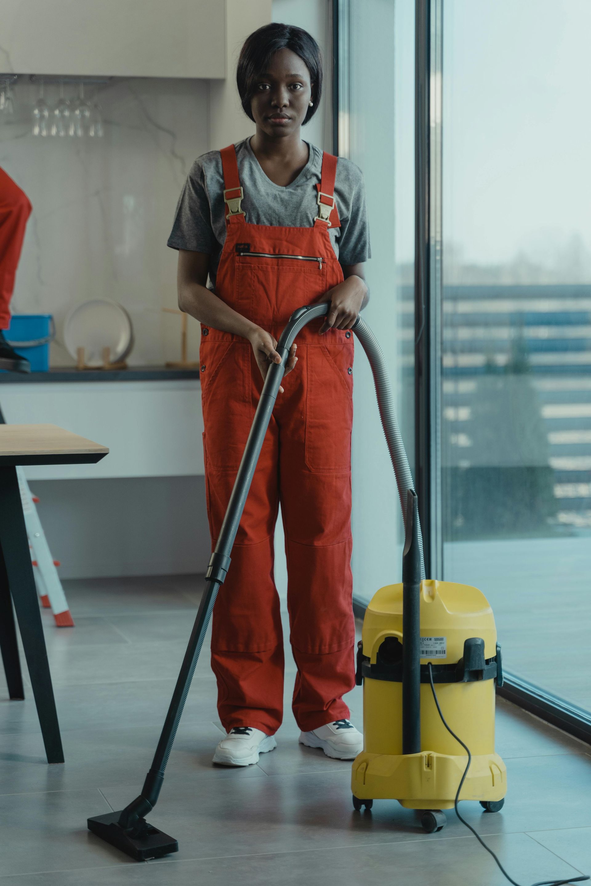 Woman in red overalls vacuums a floor in a light-filled room.