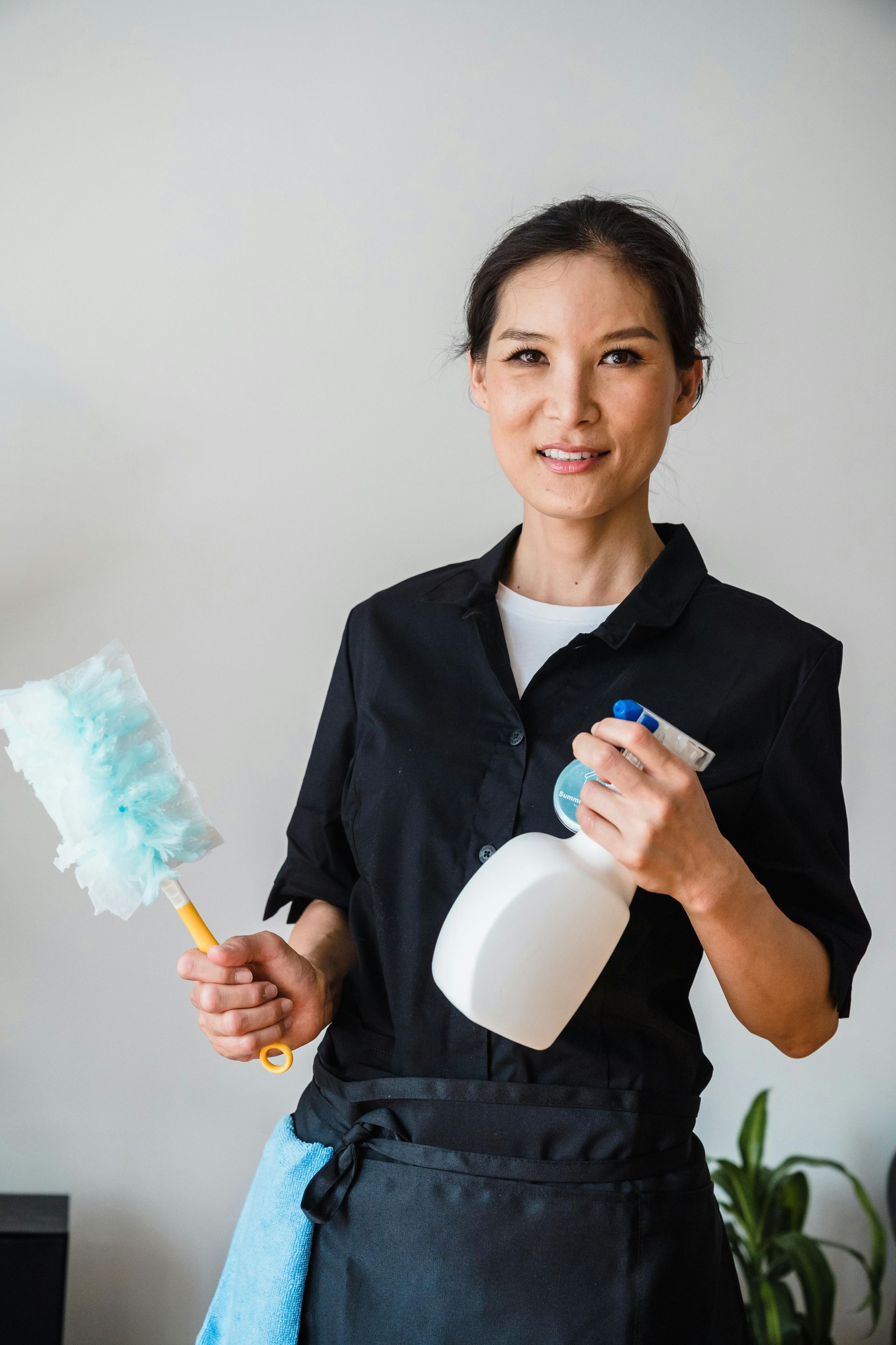Woman holding a duster and spray bottle, smiling, wearing a black uniform, indoors.