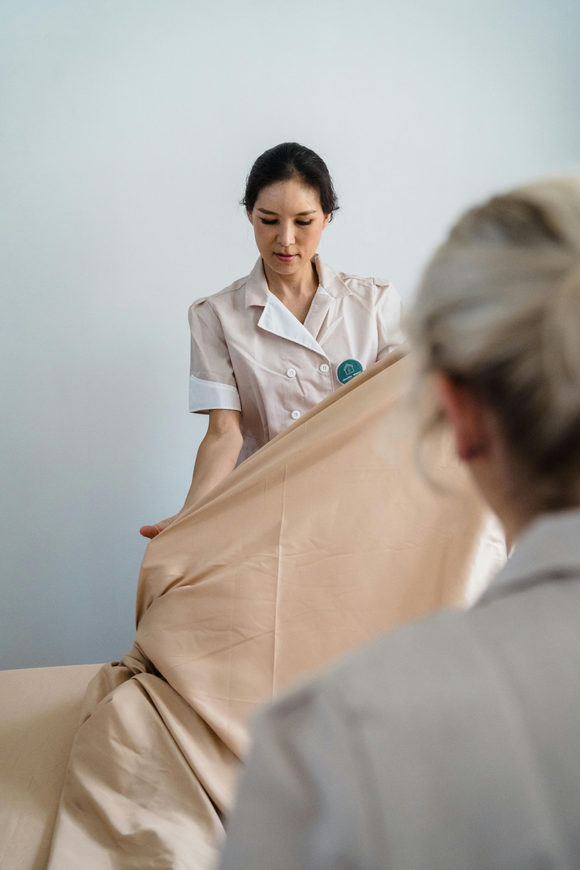 Two people making a bed with a tan sheet in a light-colored room.