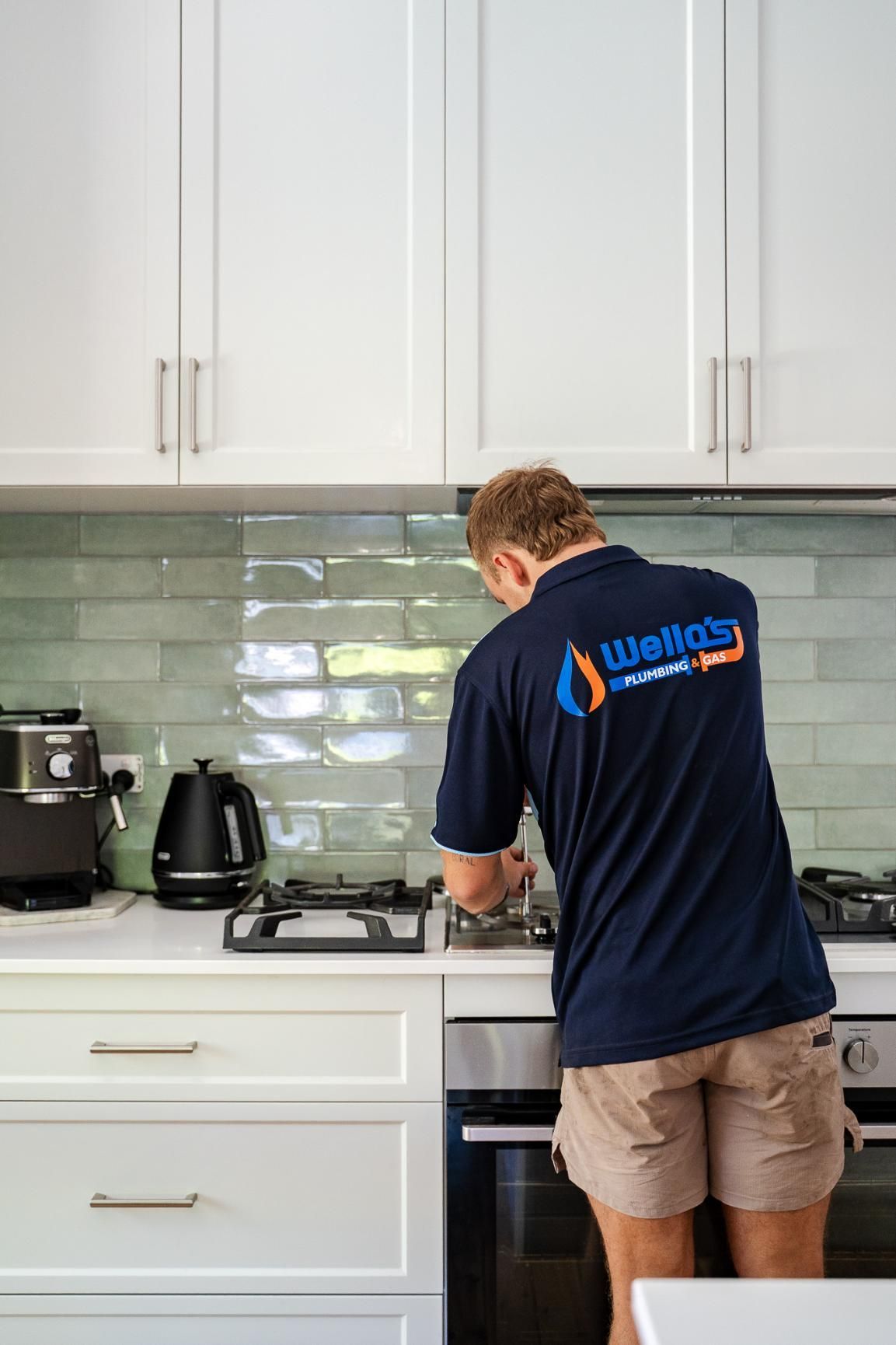 A Man in a Blue Shirt is Working on a Stove in a Kitchen — Wello's Plumbing & Gas In Thurgoona, NSW
