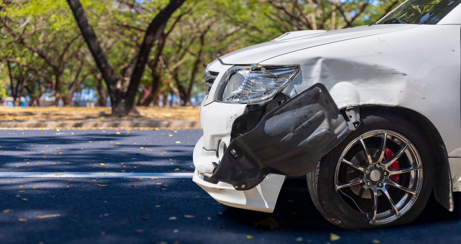 A damaged white car with a hanging front bumper panel, parked on an asphalt road near trees.