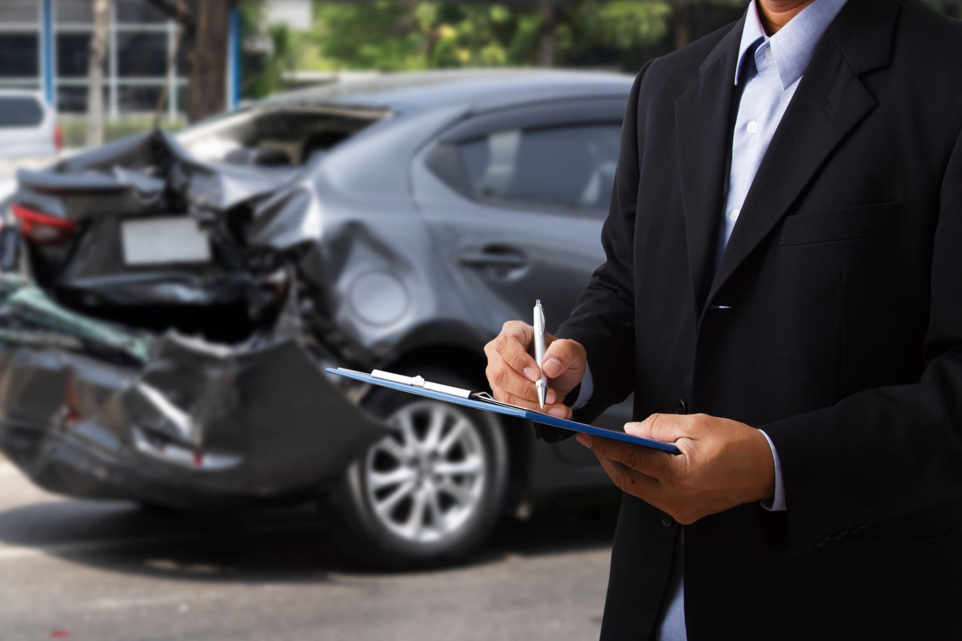 A person in a suit writes on a clipboard in front of a dark car with significant rear-end damage.