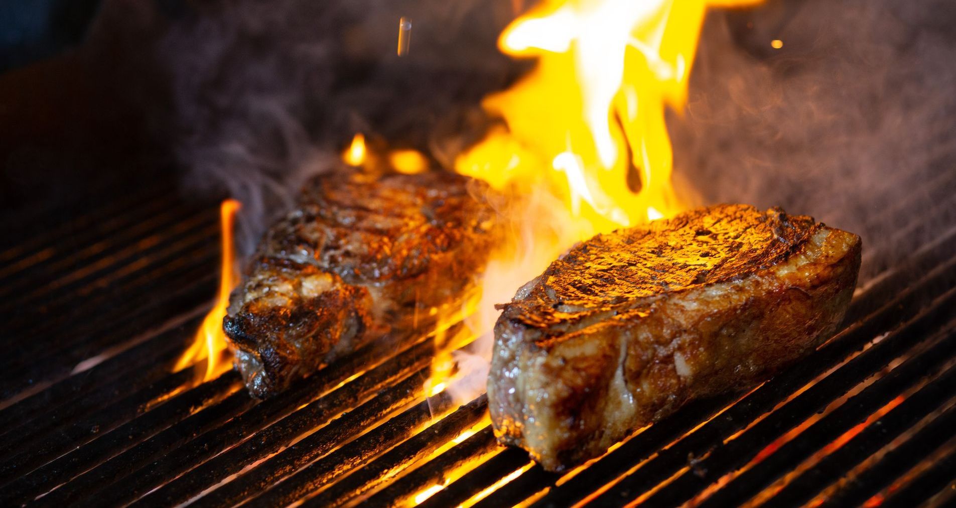 Dos filetes se están cocinando en una parrilla con llamas saliendo de ellos.