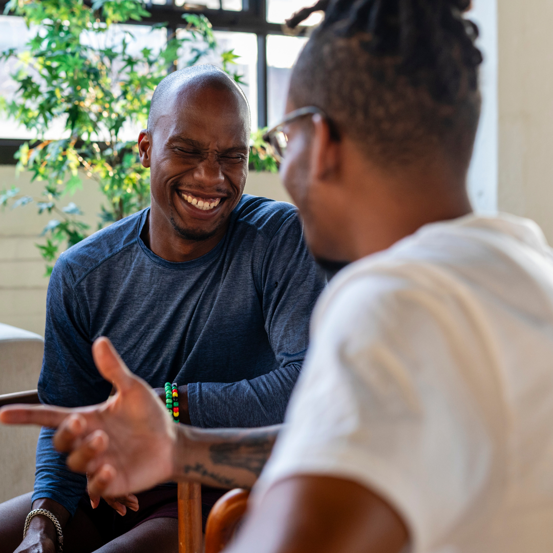 Two Black men smiling and talking in a bright room; one points his finger and gestures, the other laughs.
