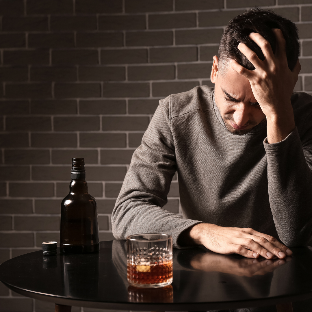 A man with his head in his hand sits at a table with a bottle of liquor and a drink, appearing distressed.