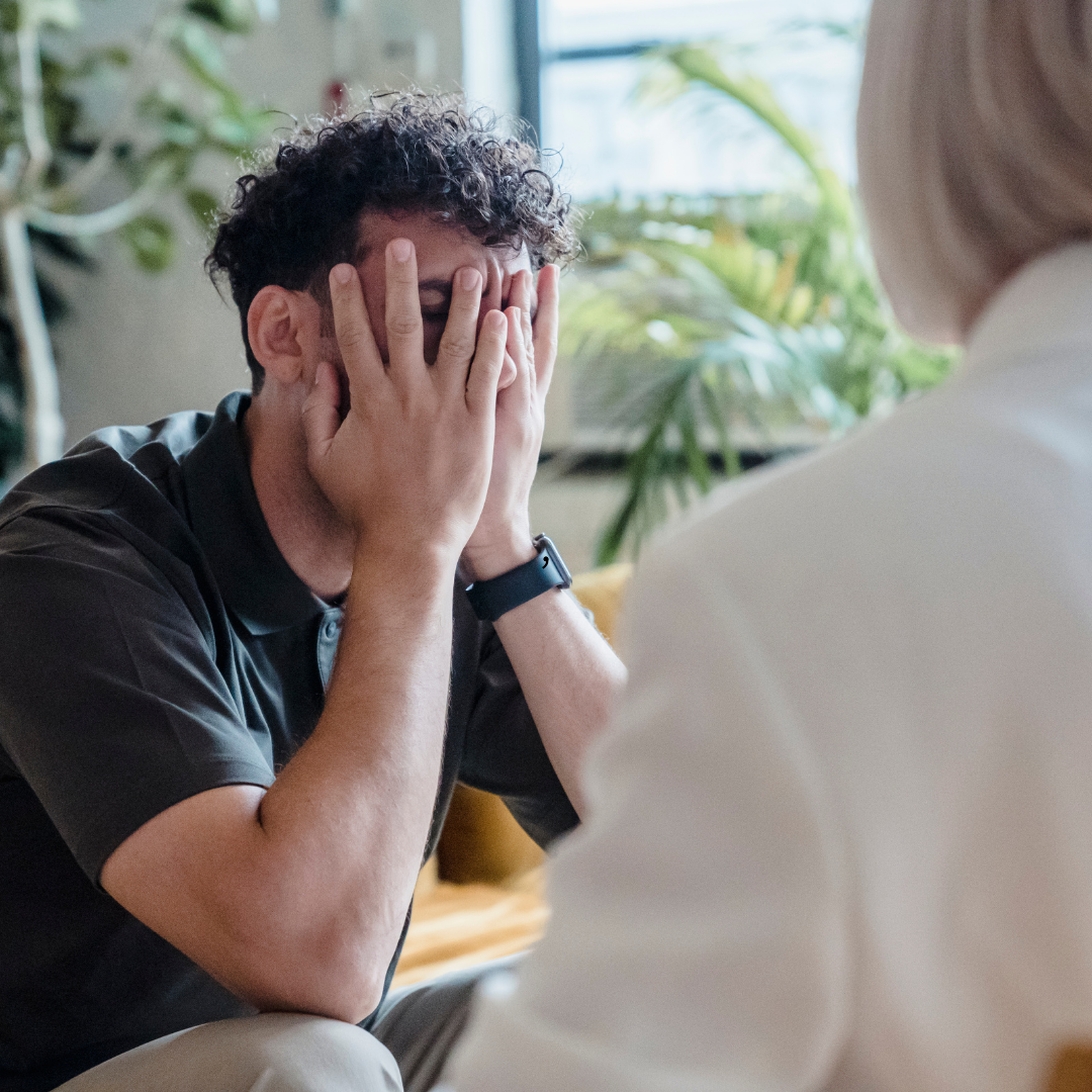 Man with curly hair covering his face, sitting in front of a person, likely in a therapy session.