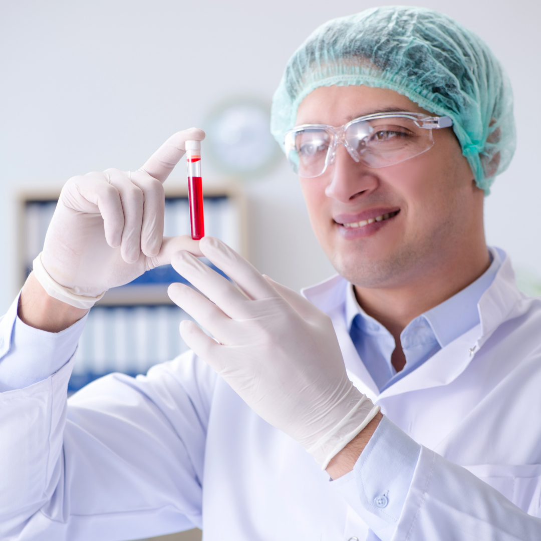 A lab technician in a white coat, gloves, and safety glasses smiles while holding a vial of red liquid, possibly blood.