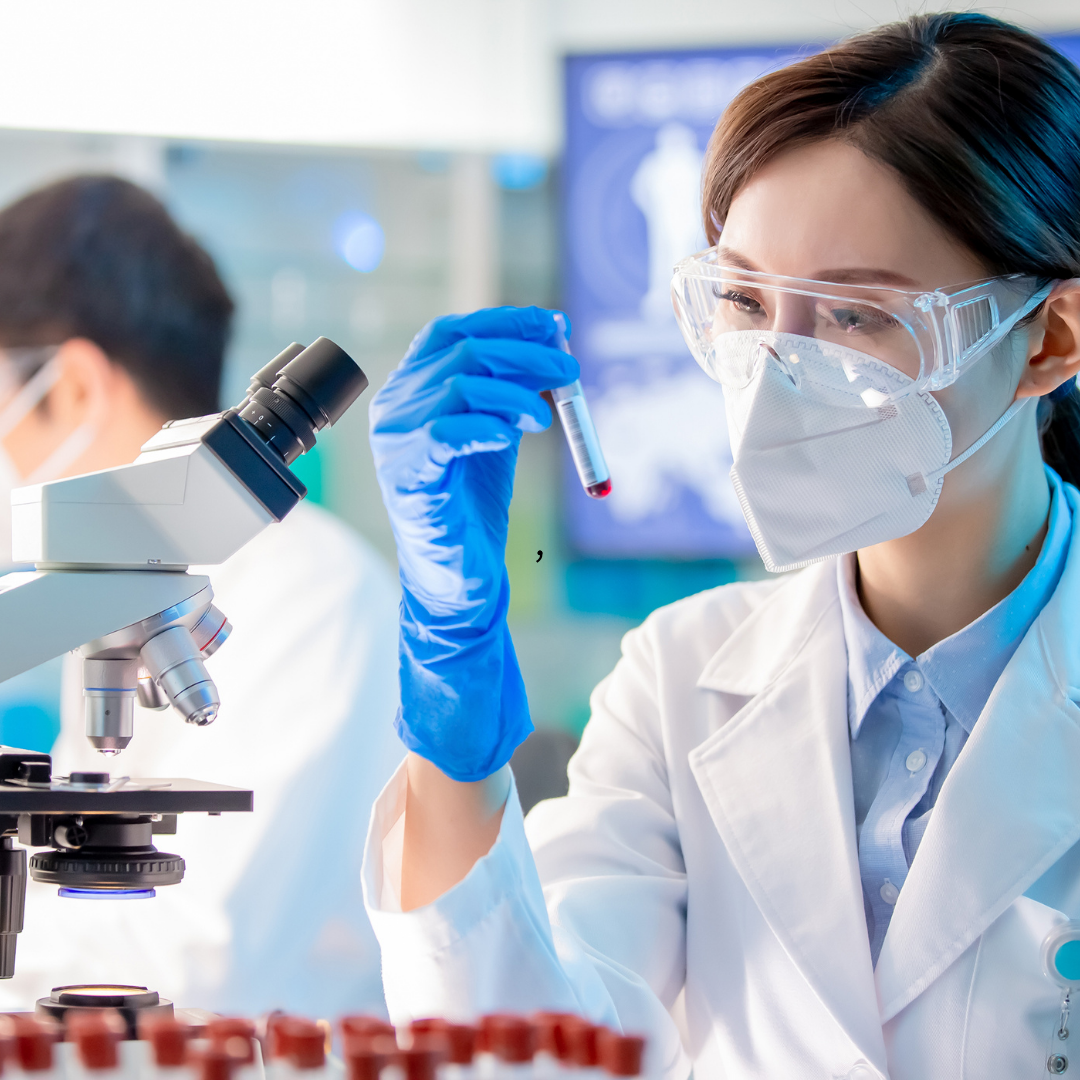 A lab worker in safety glasses and mask holds a test tube of blood, with a microscope and another worker visible.
