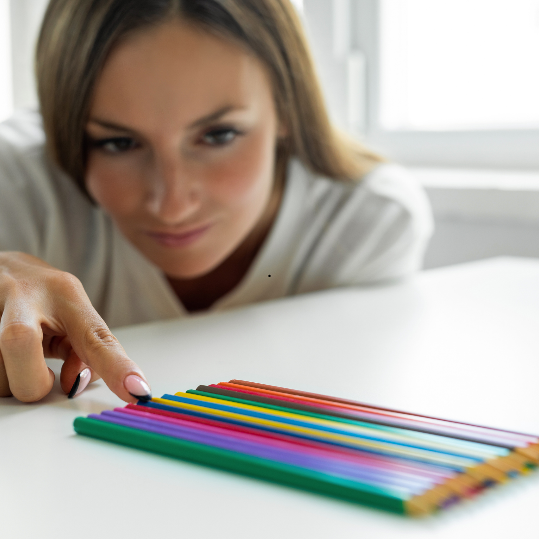 Woman pointing at a row of colored pencils on a white table. She is leaning forward, focused expression.