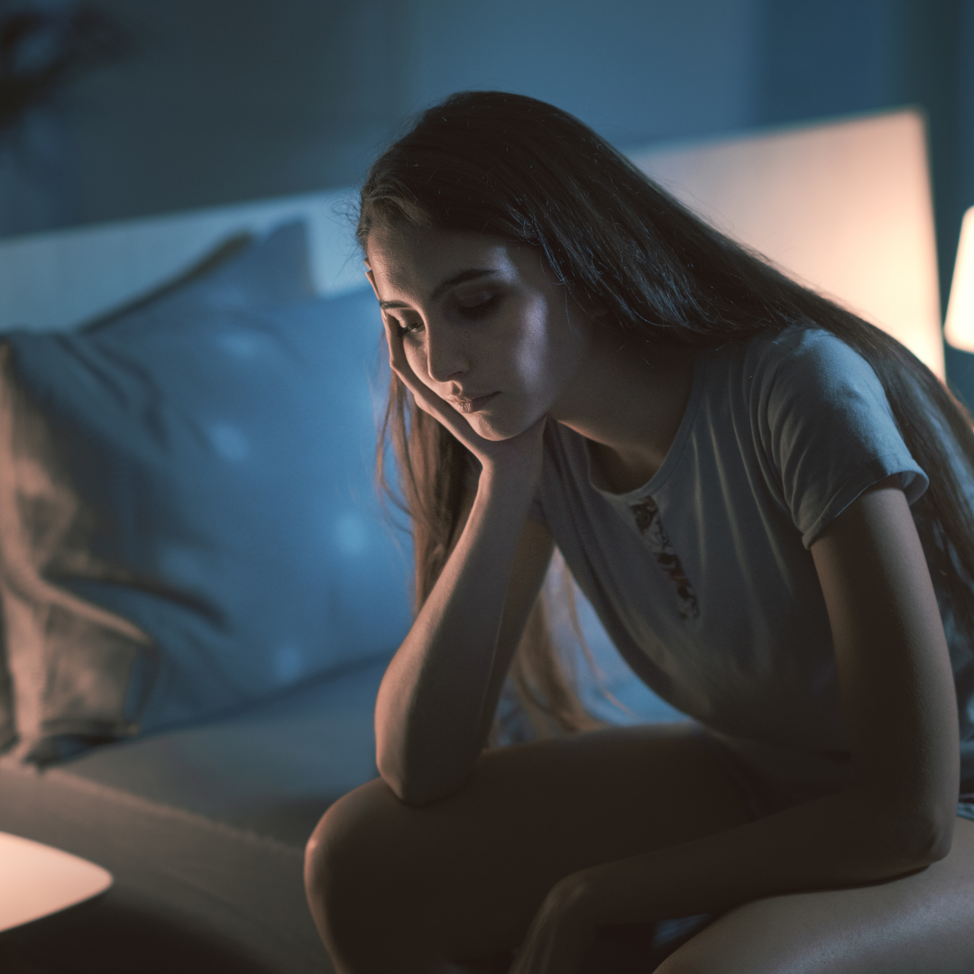 Woman sitting on a bed at night, looking downcast, with a hand on her cheek. Dim room, soft lighting.