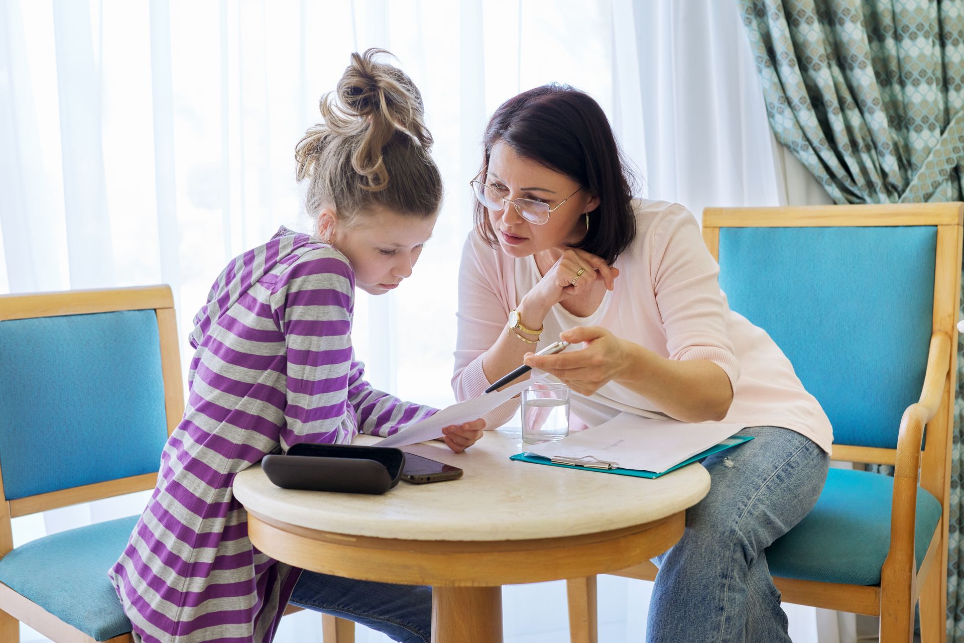 Woman and girl sit at a table looking at papers; woman points with pen, appearing to instruct.