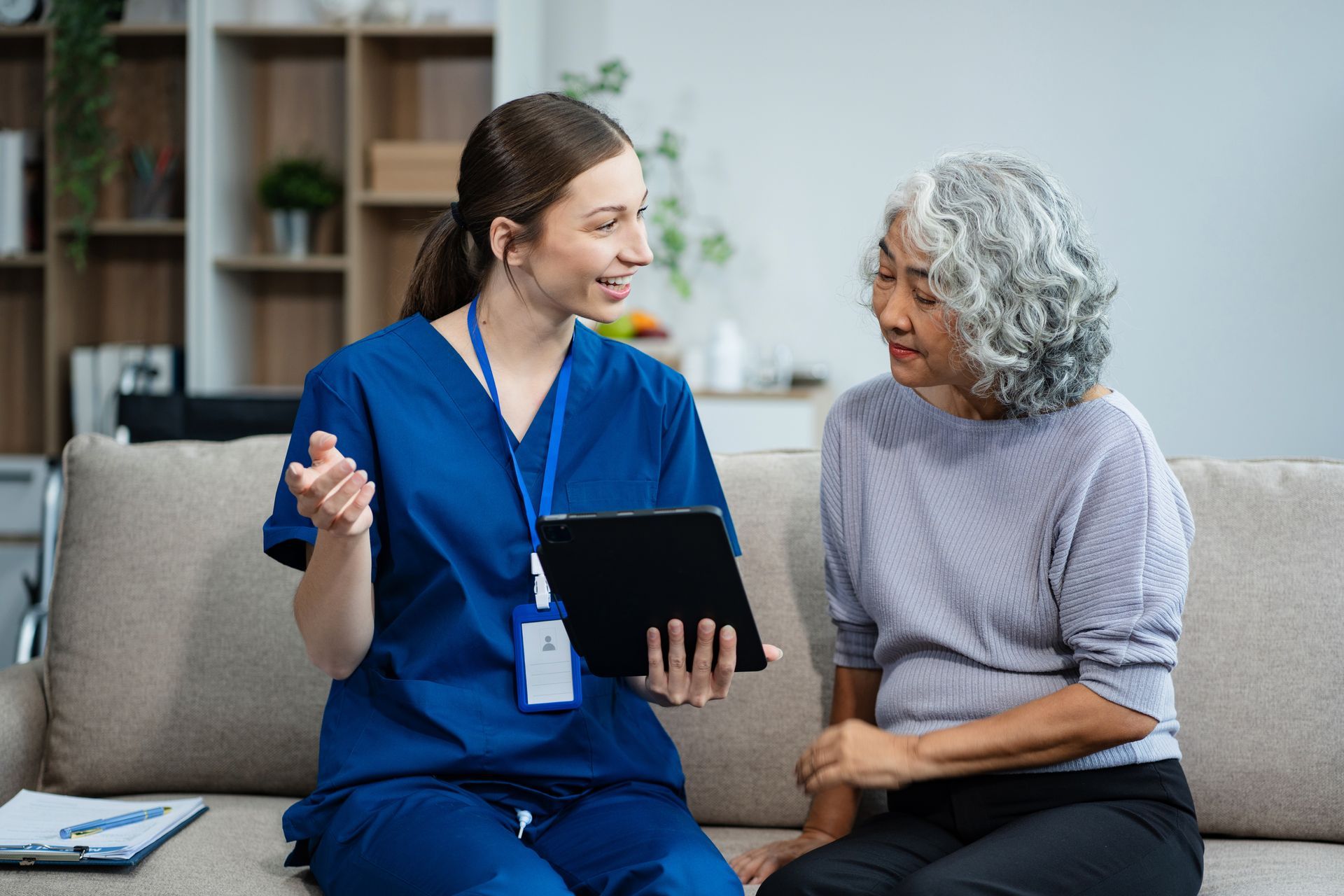 A healthcare worker in blue scrubs consults with an older woman on a sofa, both smiling. They are in a home setting.