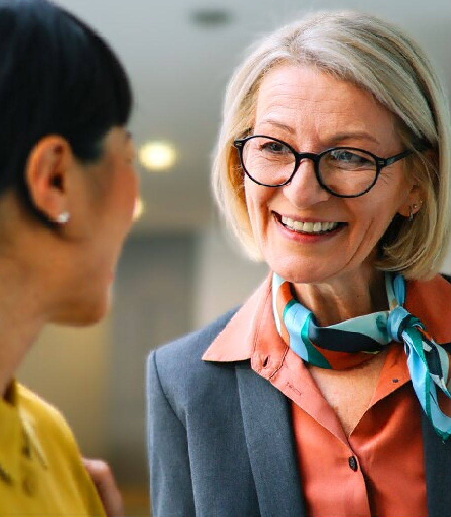 A woman wearing glasses and a scarf is smiling while talking to another woman.