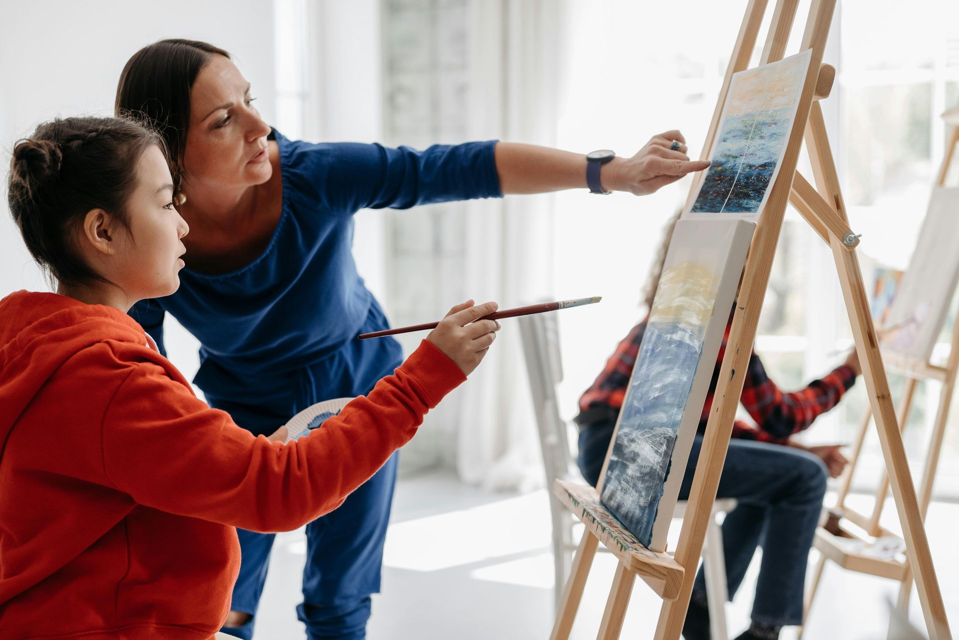 A woman is teaching a young girl how to paint on an easel.