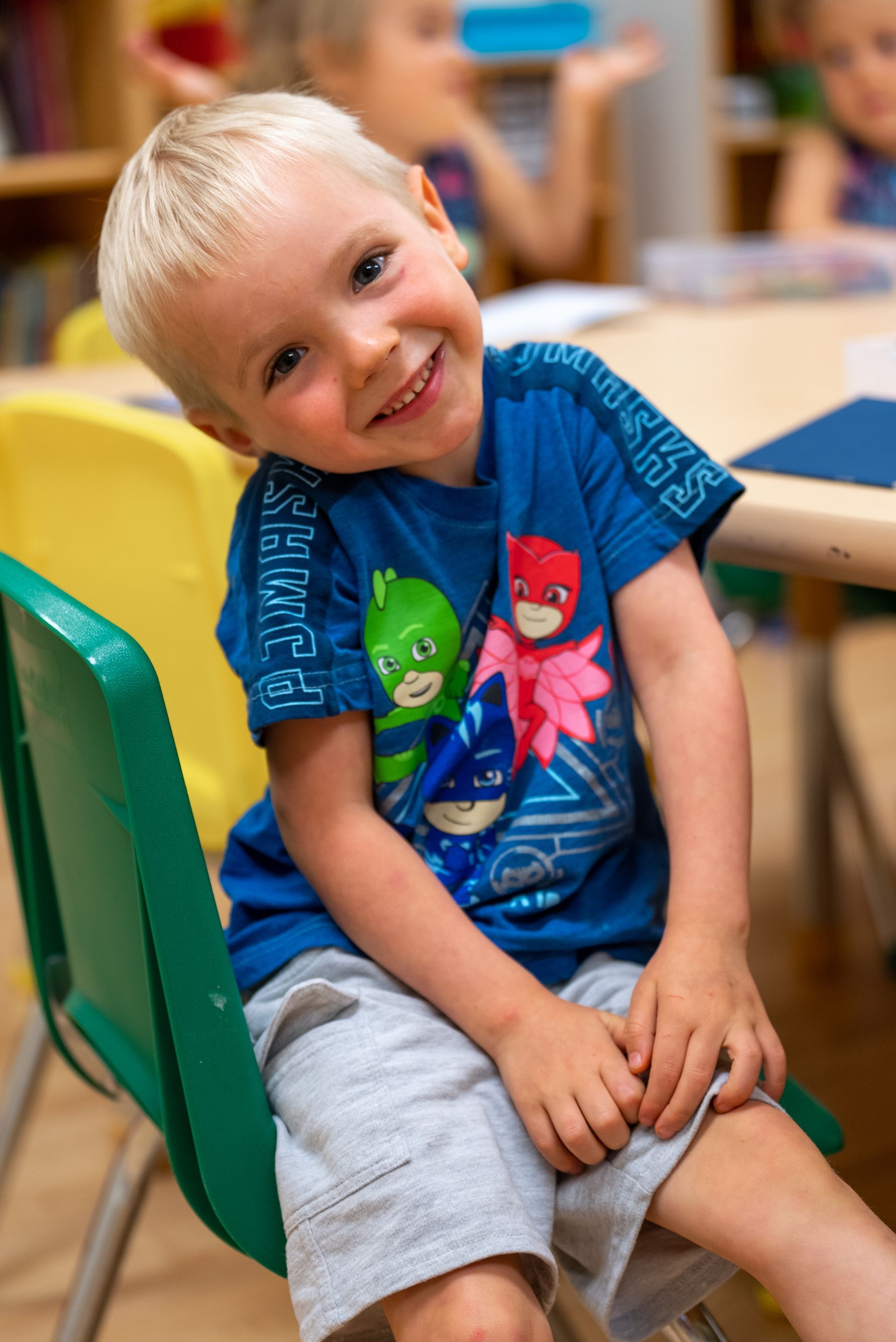 A young boy wearing a pj masks shirt is sitting in a green chair.