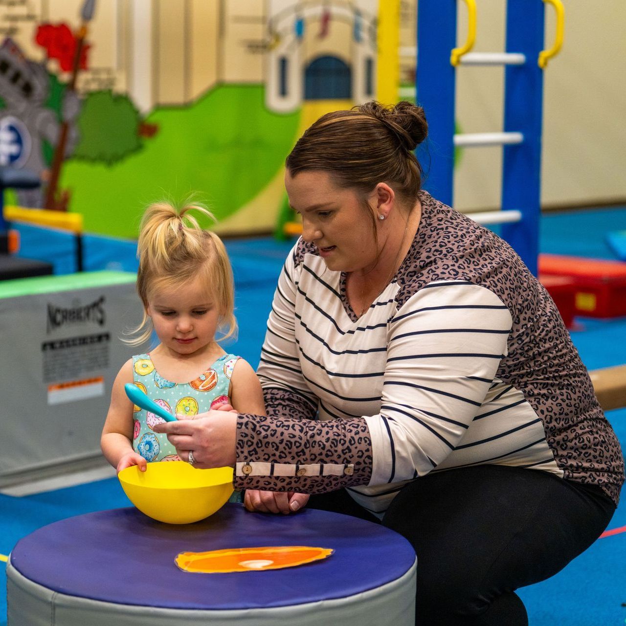 A woman is kneeling down next to a little girl playing with a yellow bowl.