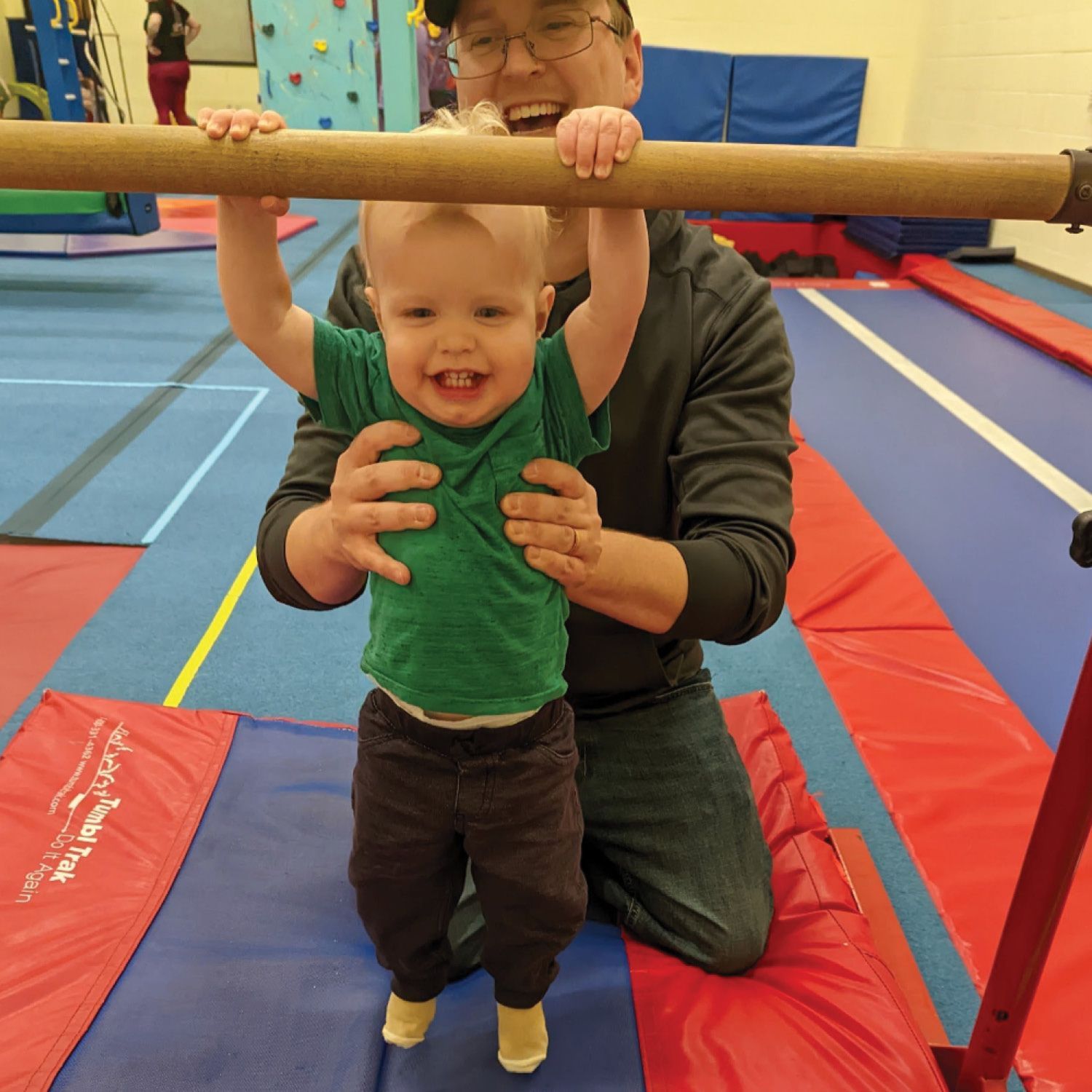 A man is kneeling next to a little boy holding a bar in a gym