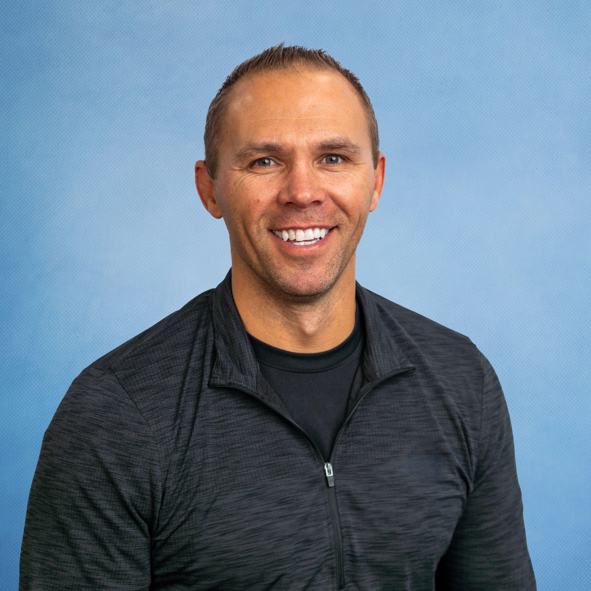 A man wearing a black shirt and a grey jacket is smiling for the camera.