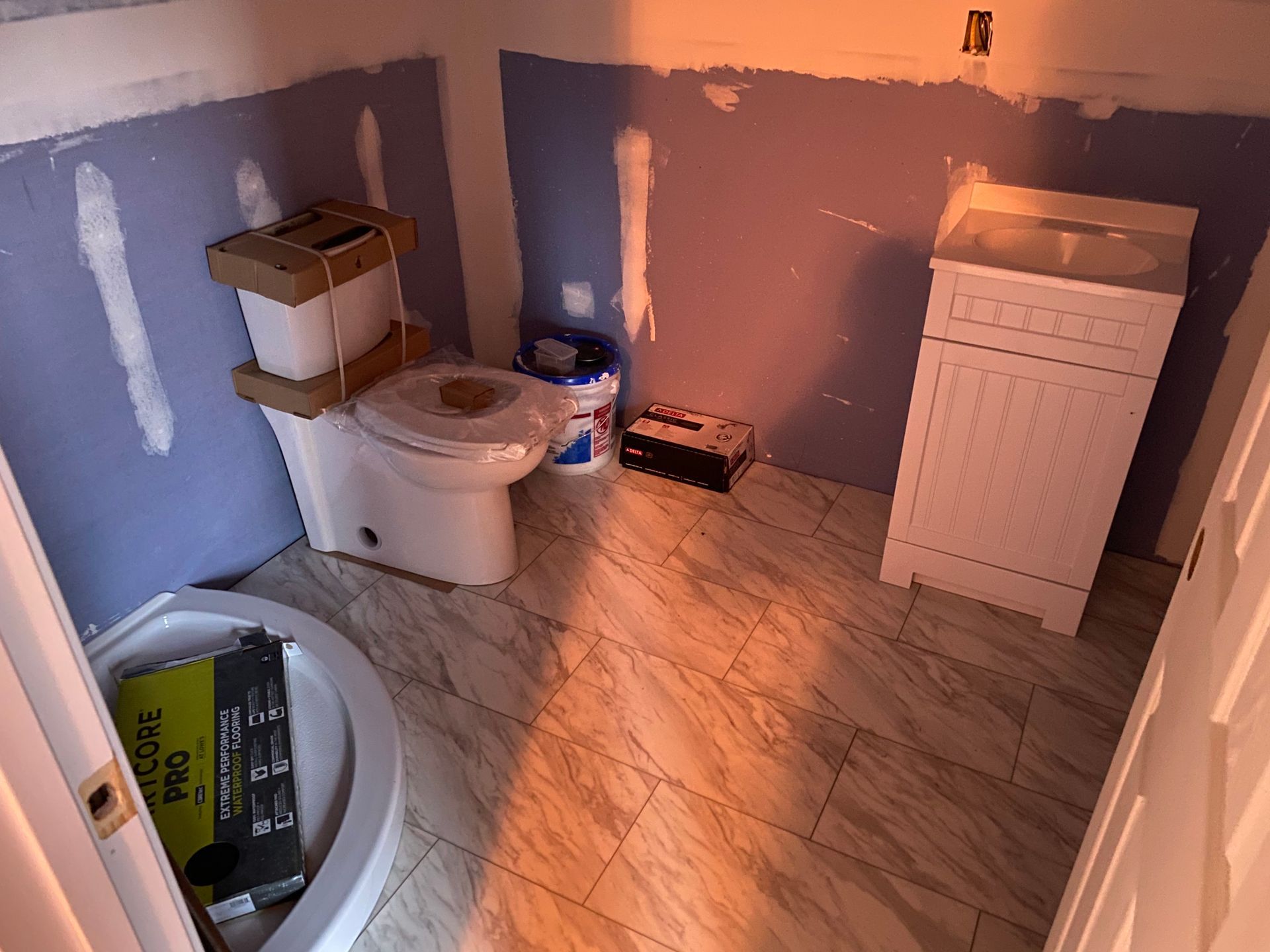 Bathroom under renovation, with new toilet, vanity, and tile floor. Blue walls, sunlight.