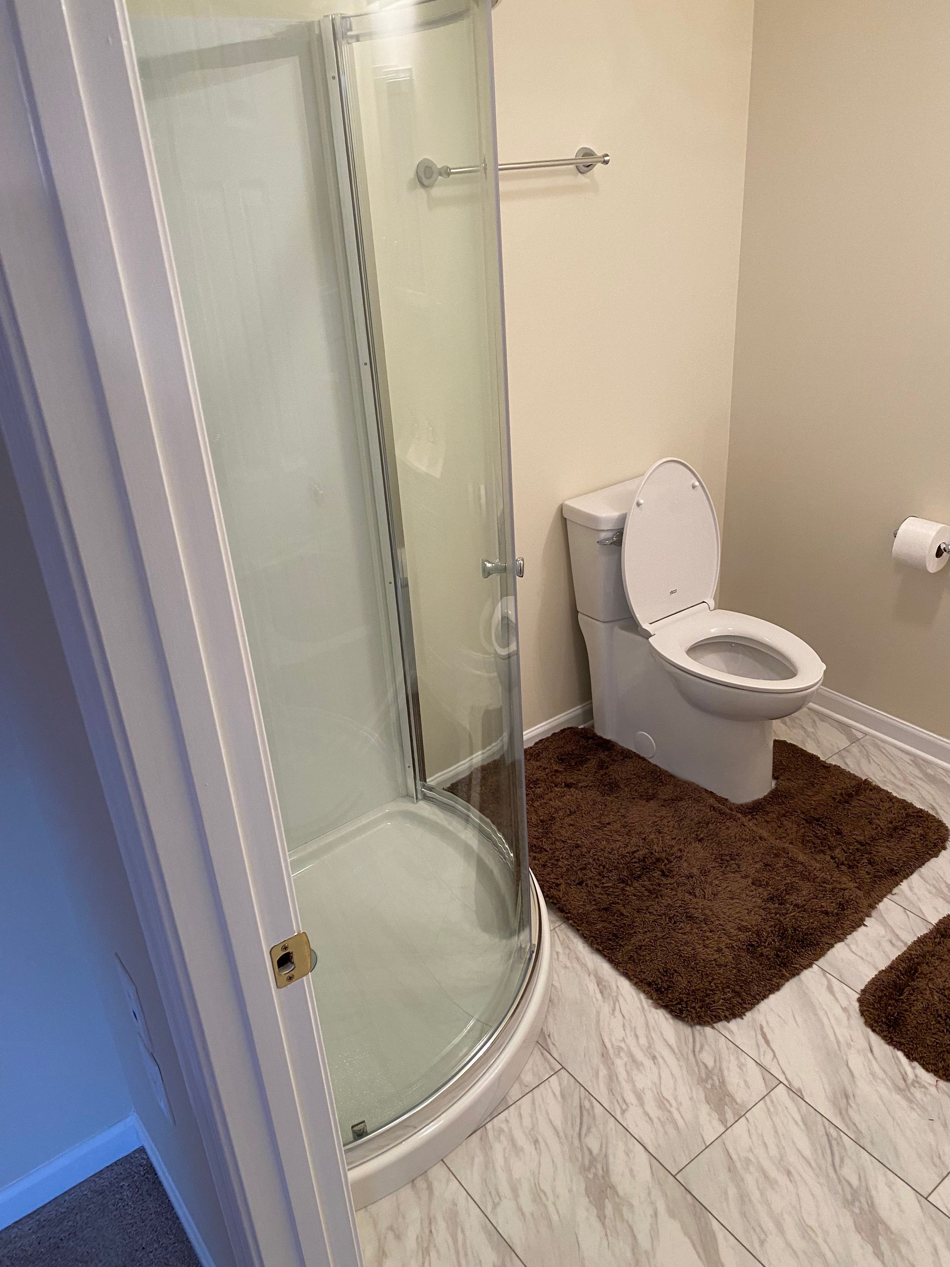 Bathroom with curved shower, toilet, and brown rugs on light-colored tiled floor.