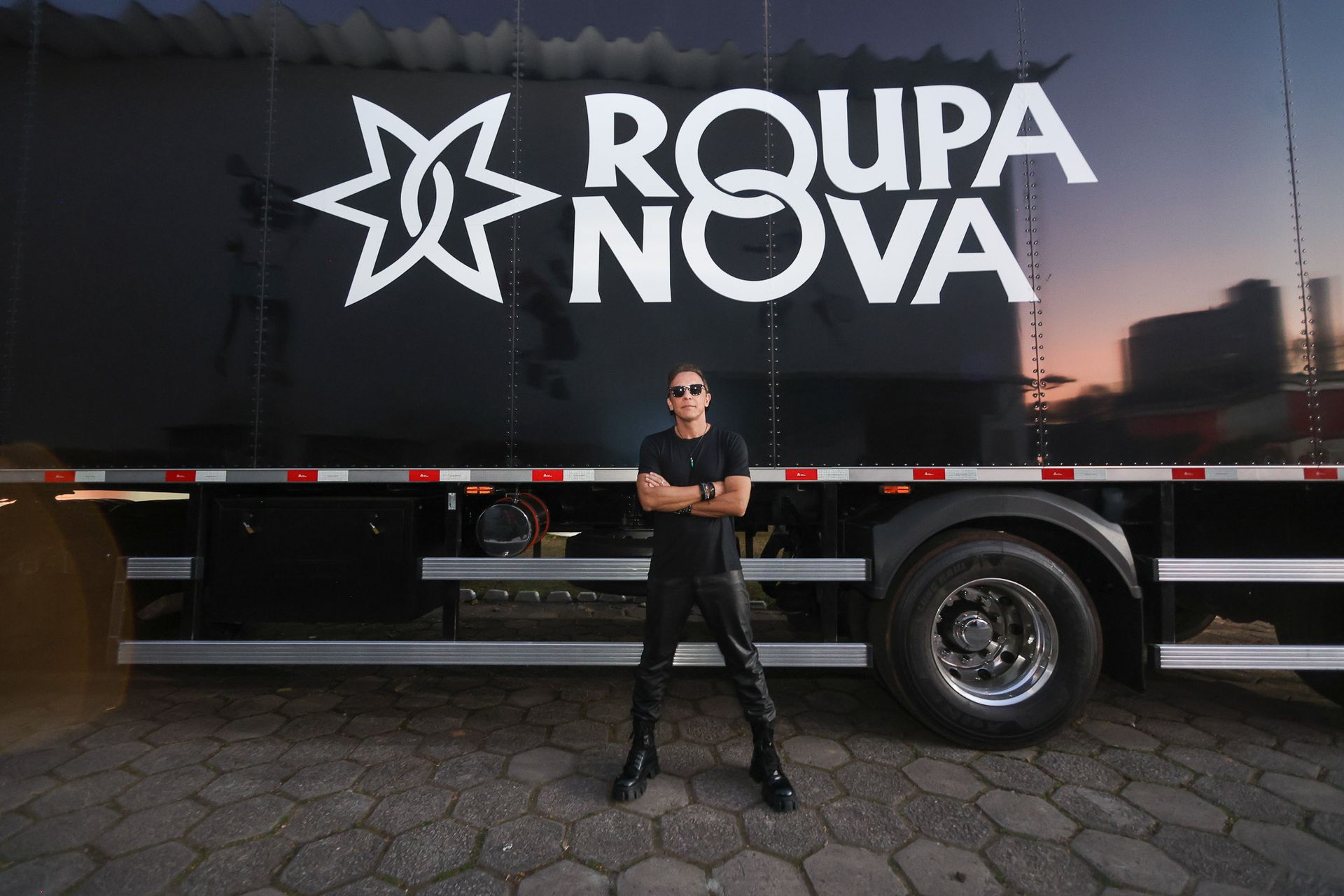 Um artista está de pé com os braços cruzados em frente a um grande caminhão preto com a marca Roupa Nova.
