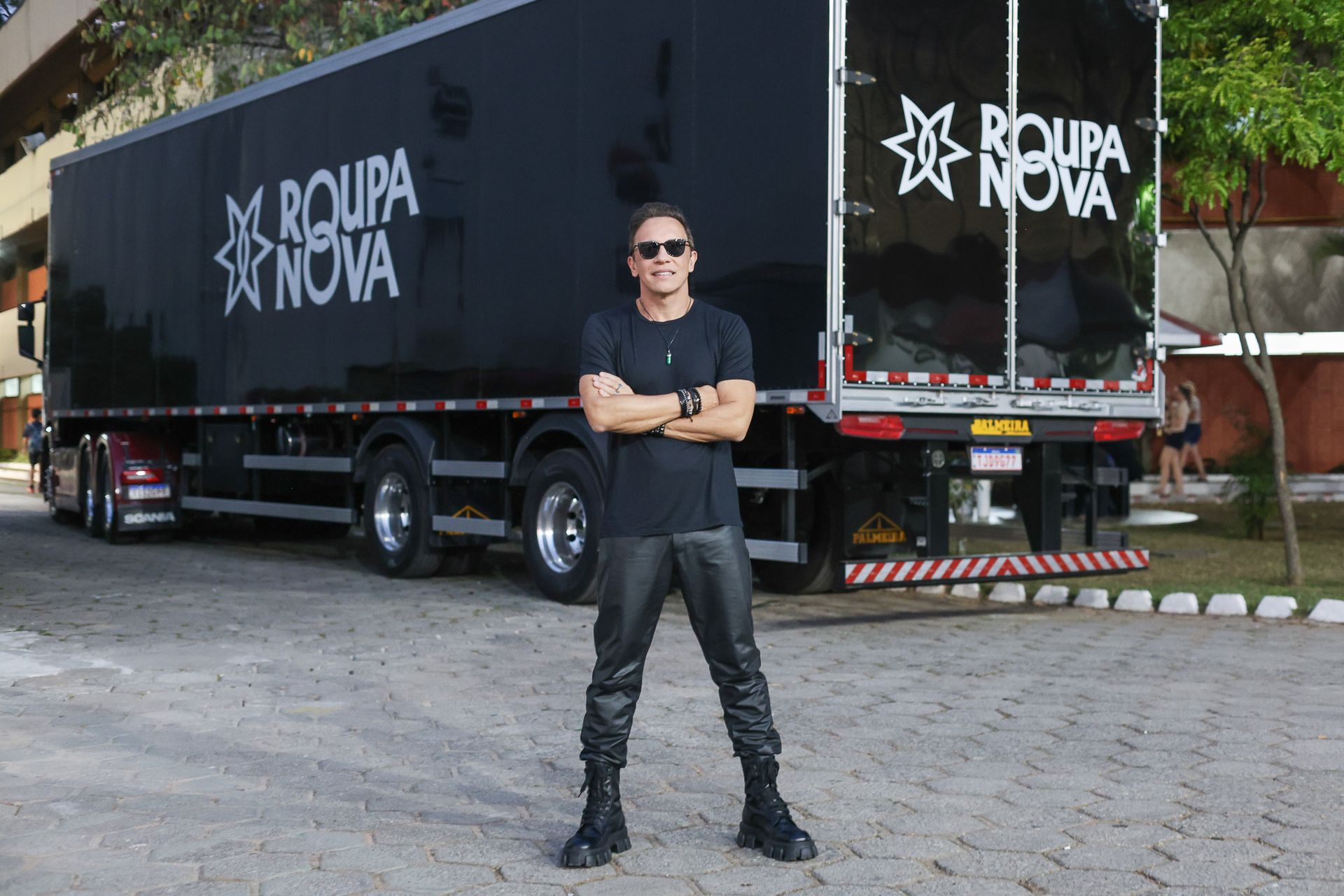 Um artista de camisa preta, calça de couro e óculos escuros está em frente a um caminhão preto da turnê Roupa Nova.