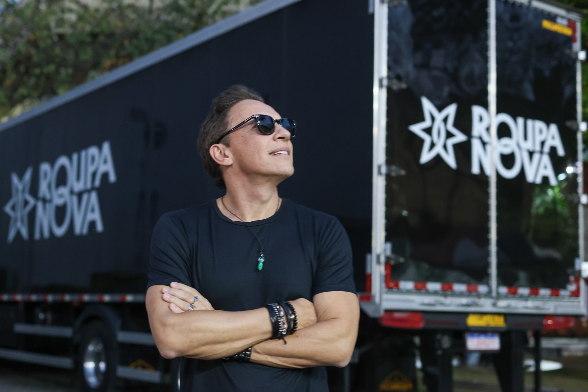 Um artista de óculos escuros e braços cruzados olhando para cima em frente a um caminhão com a marca Roupa Nova.