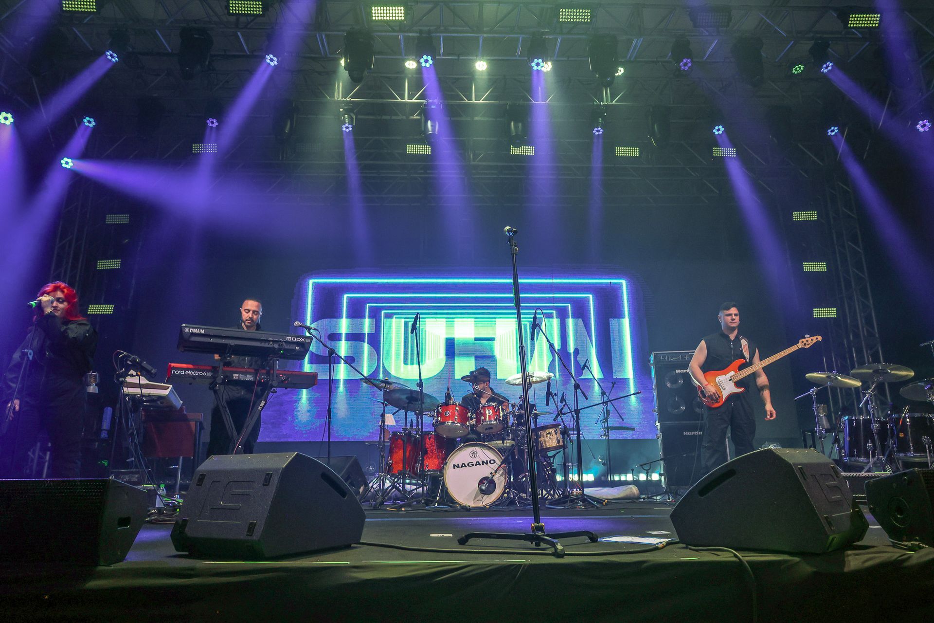 Uma banda se apresentando em um palco de shows com iluminação roxa, um painel de neon ao fundo e monitores de palco.