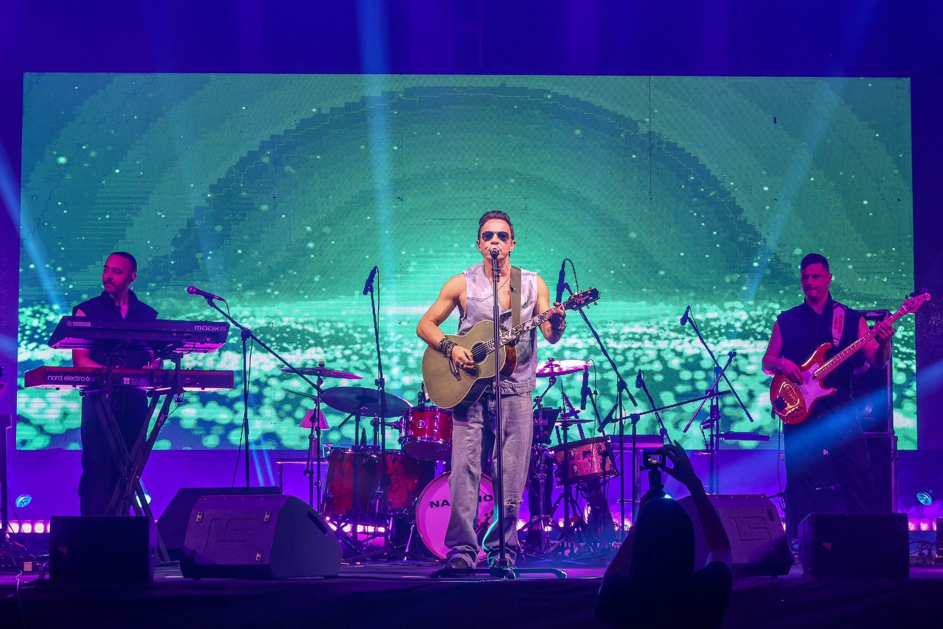 Uma banda musical se apresenta em um palco com iluminação azul, incluindo um vocalista com guitarra e dois instrumentistas.