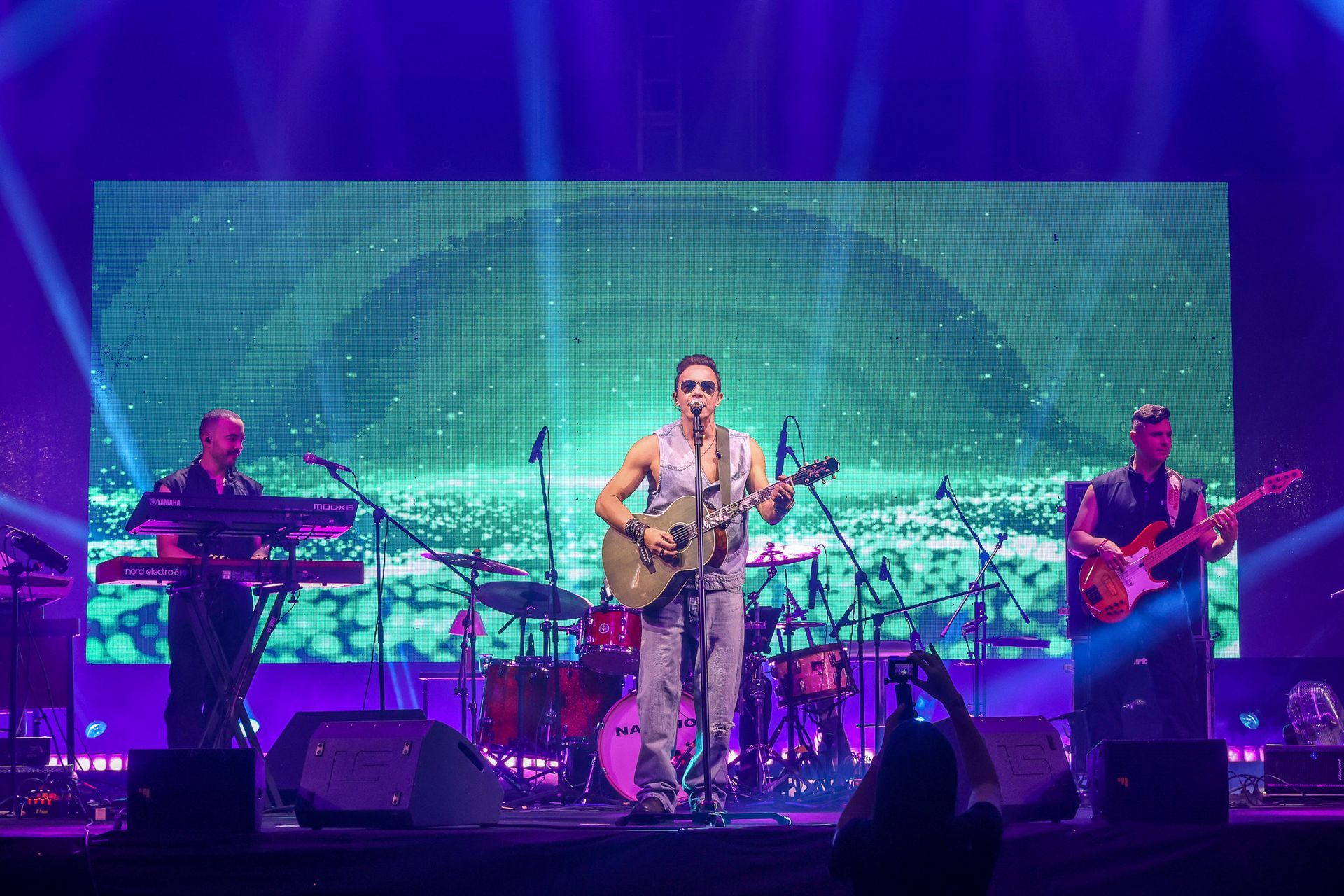 Uma banda se apresenta em um palco de shows com iluminação roxa e um telão digital brilhante com um arco verde.
