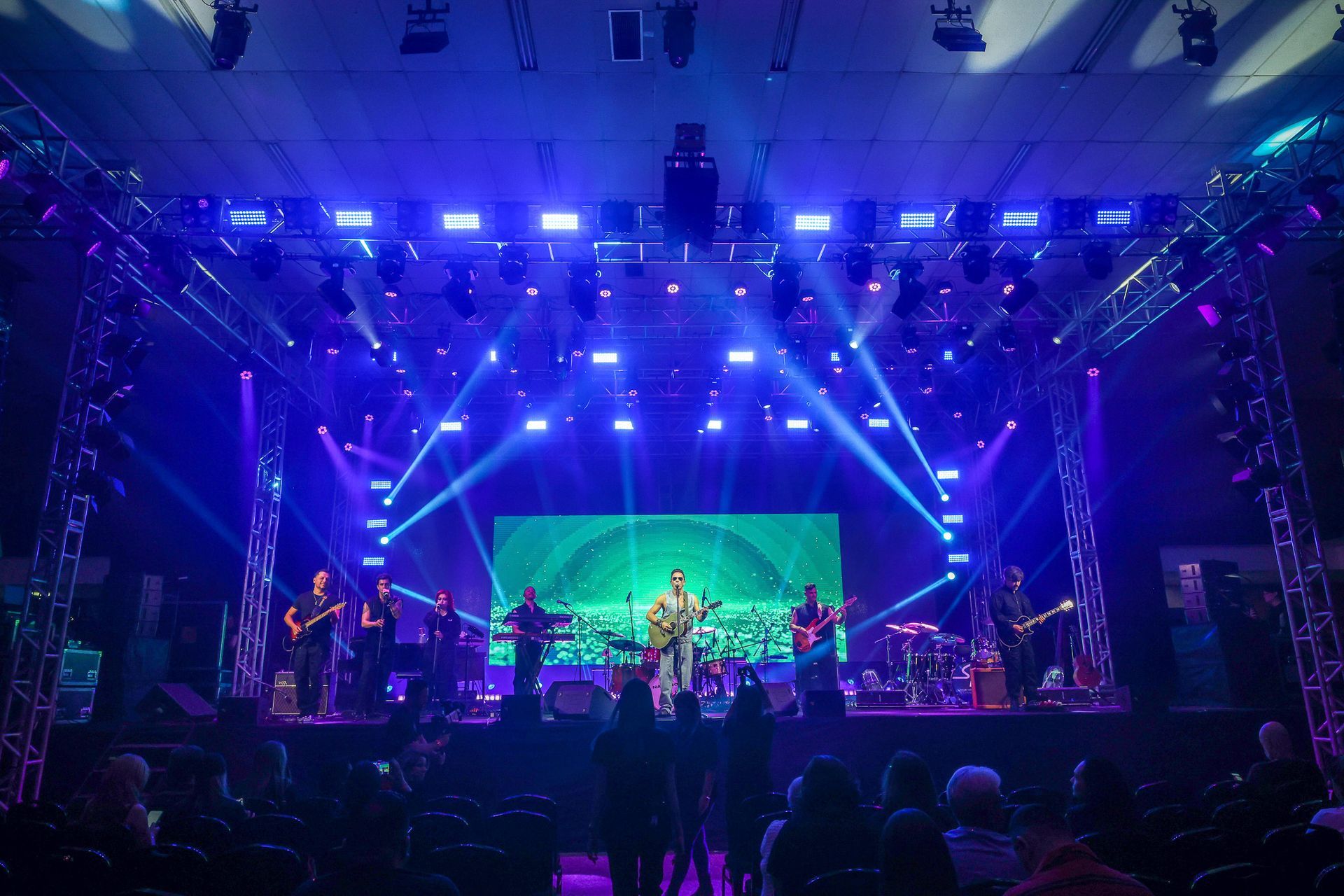 Uma banda ao vivo se apresenta em um palco de concertos iluminado por holofotes azuis brilhantes, assistida por uma plateia.