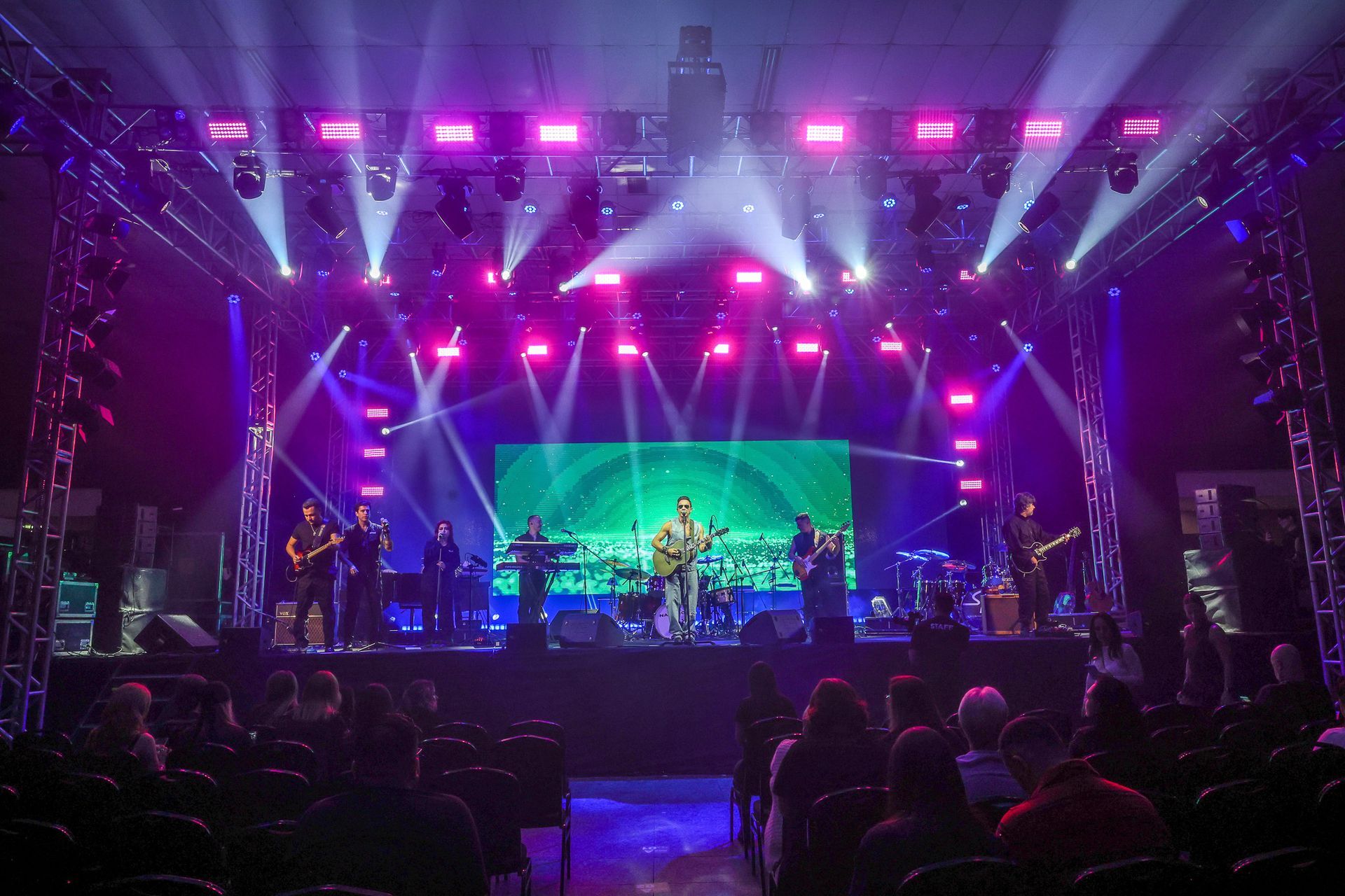 Uma banda se apresenta em um palco de shows bem iluminado com holofotes roxos e brancos, vista da perspectiva da plateia.