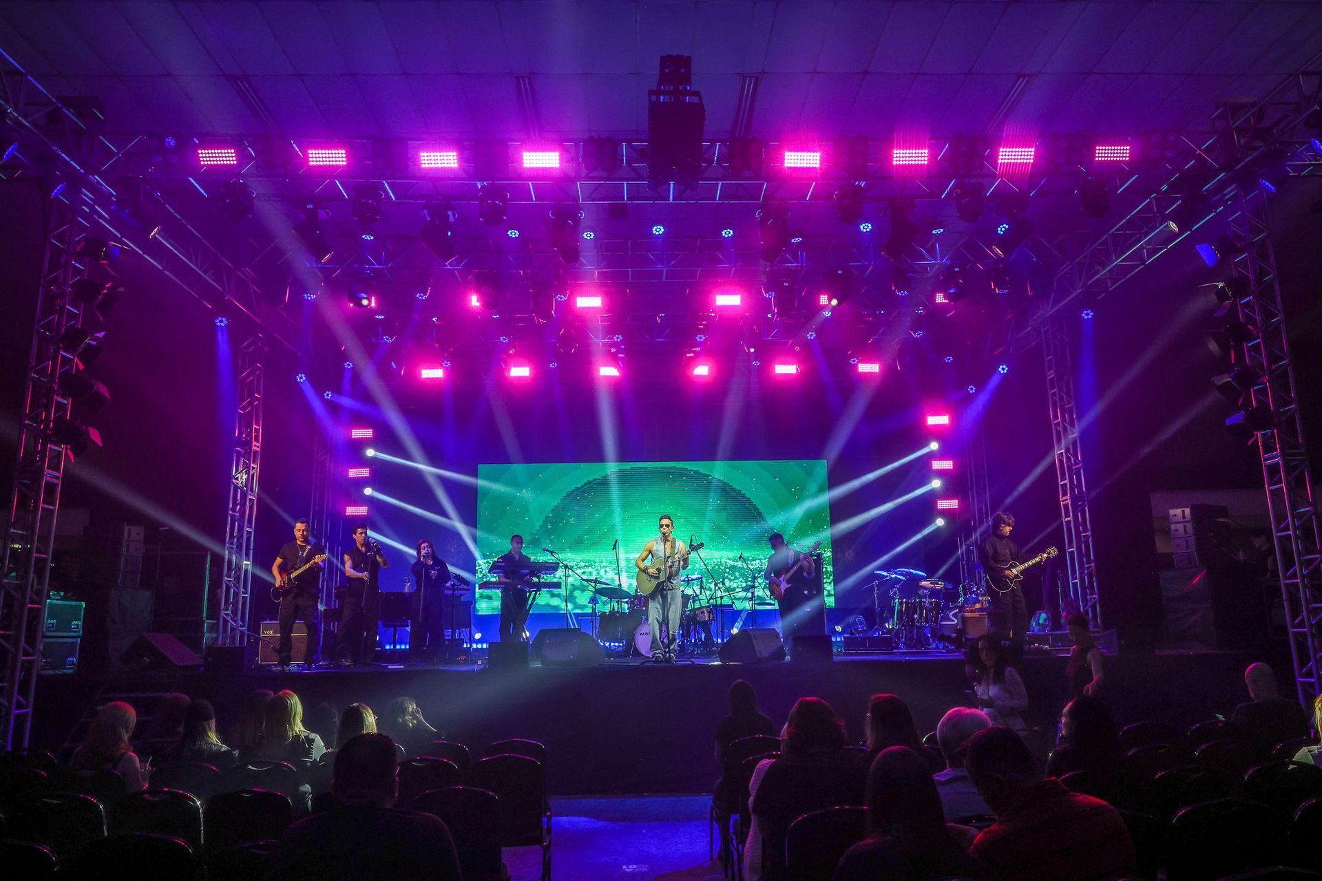 Uma banda se apresenta em um palco de shows bem iluminado com luzes roxas e um telão ao fundo, vista da plateia.
