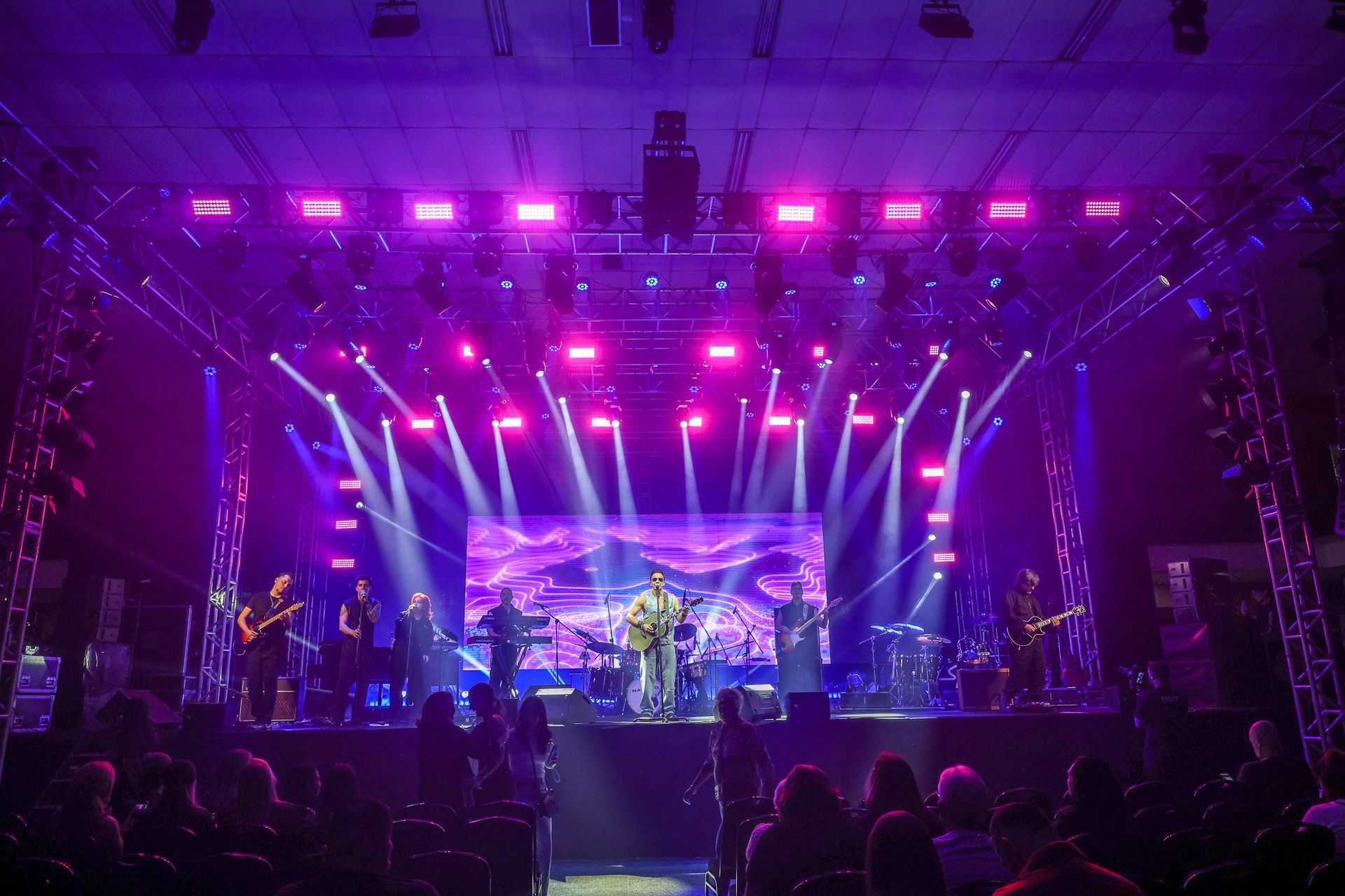 Uma banda musical se apresenta em um palco banhado por luzes brilhantes em tons de roxo e rosa, diante de uma plateia.