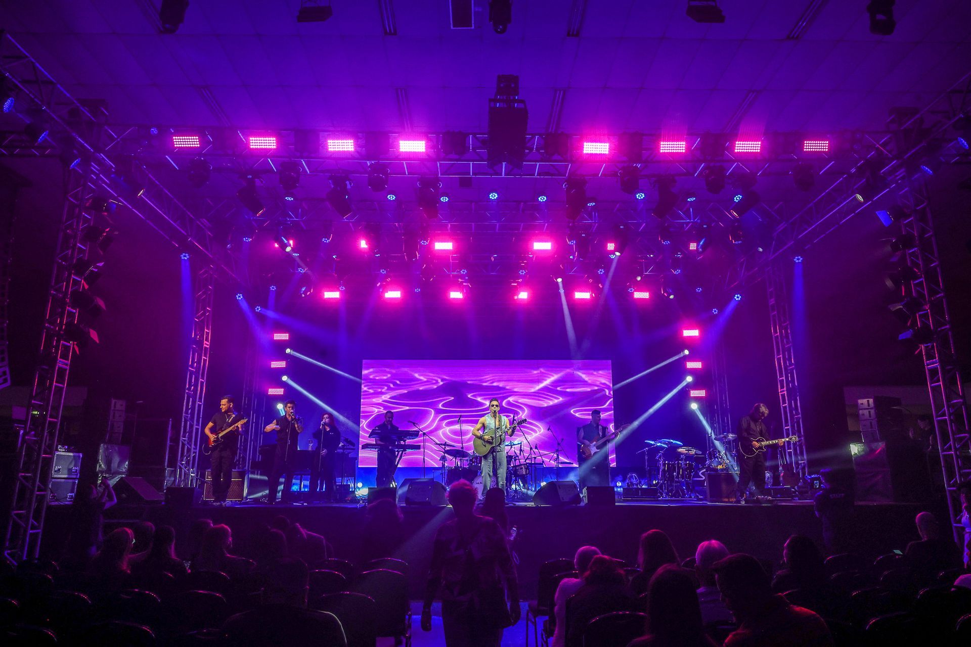 Uma banda se apresenta em um palco de shows iluminado por luzes de palco rosa e roxas brilhantes diante de uma plateia.