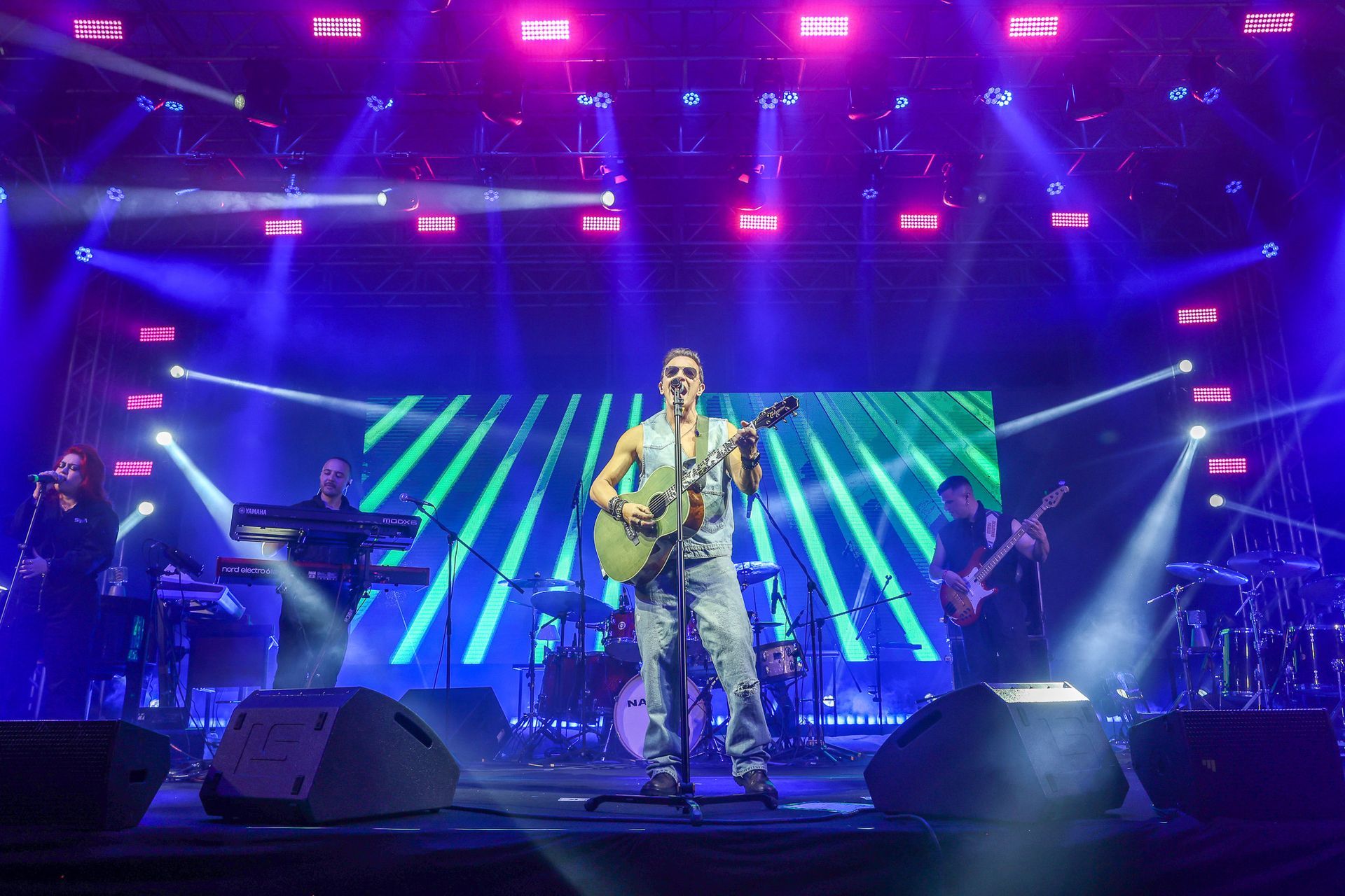 Um cantor toca violão no centro do palco, acompanhada por uma banda sob luzes vibrantes de concerto em tons de azul e roxo.