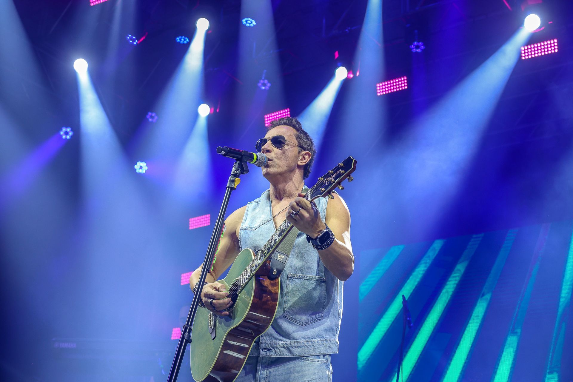 Um artista usando óculos escuros e um colete jeans toca violão no palco sob uma vibrante iluminação azul.