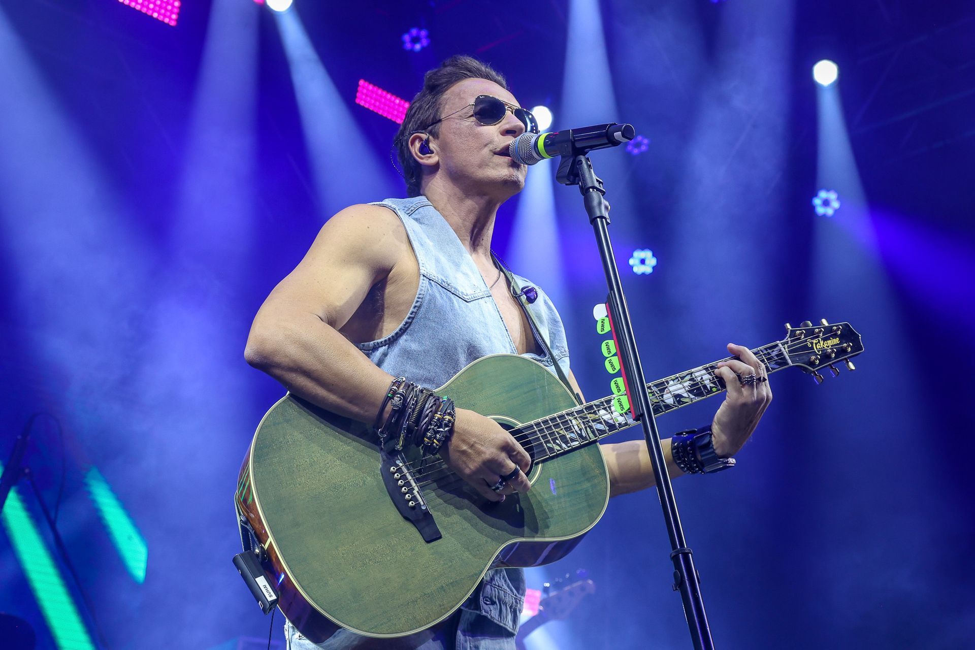 Um cantor de óculos escuros veste um colete jeans e toca um violão verde no palco sob holofotes azuis brilhantes.
