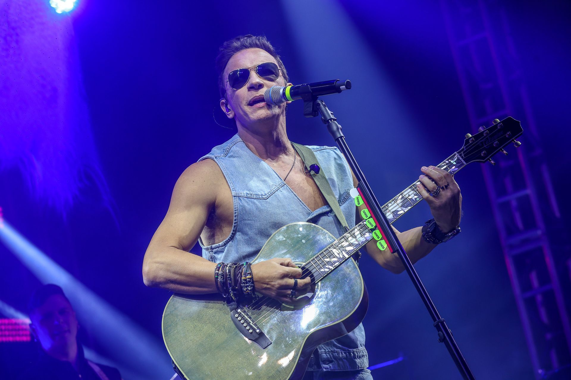 Um músico de colete jeans e óculos escuros canta ao microfone enquanto toca violão em um palco iluminado de azul.